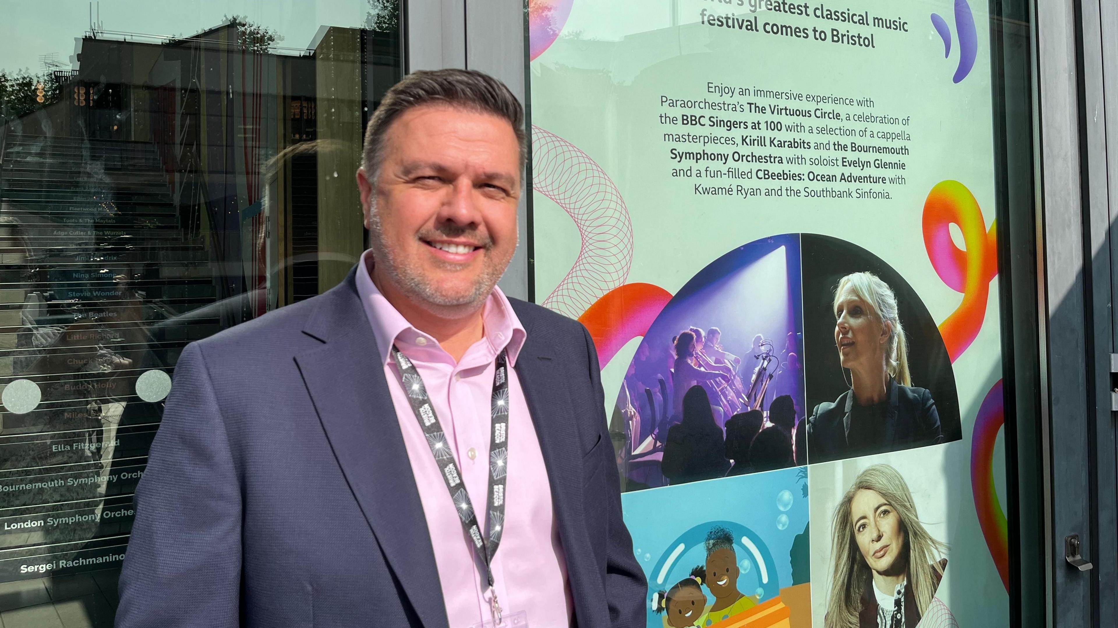 Simon Wales in navy suit with pink shirt in front of advertisement for BBC Proms at the Beacon