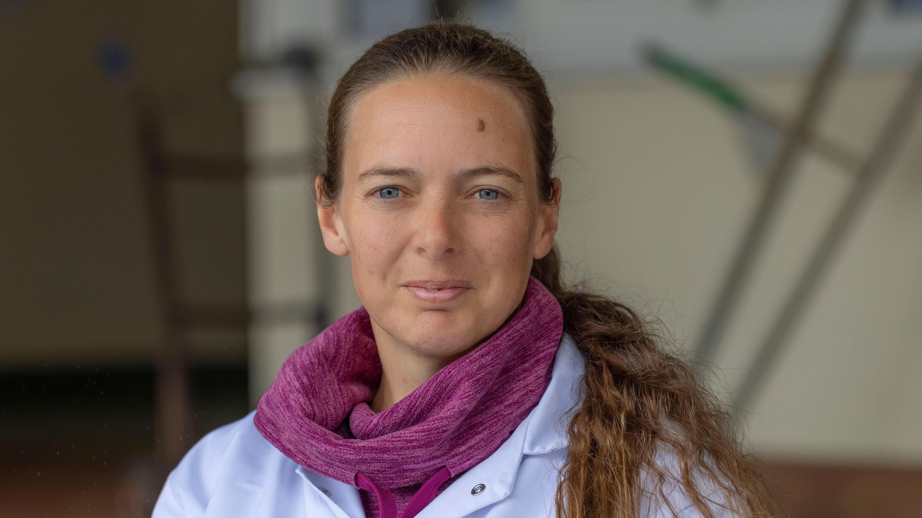 Alex Tielles standing in an area of Guernsey Dairy while wearing a white laboratory coat and a pink coloured fleece underneath. Her brown hair is in a ponytail which is draped over her left shoulder.