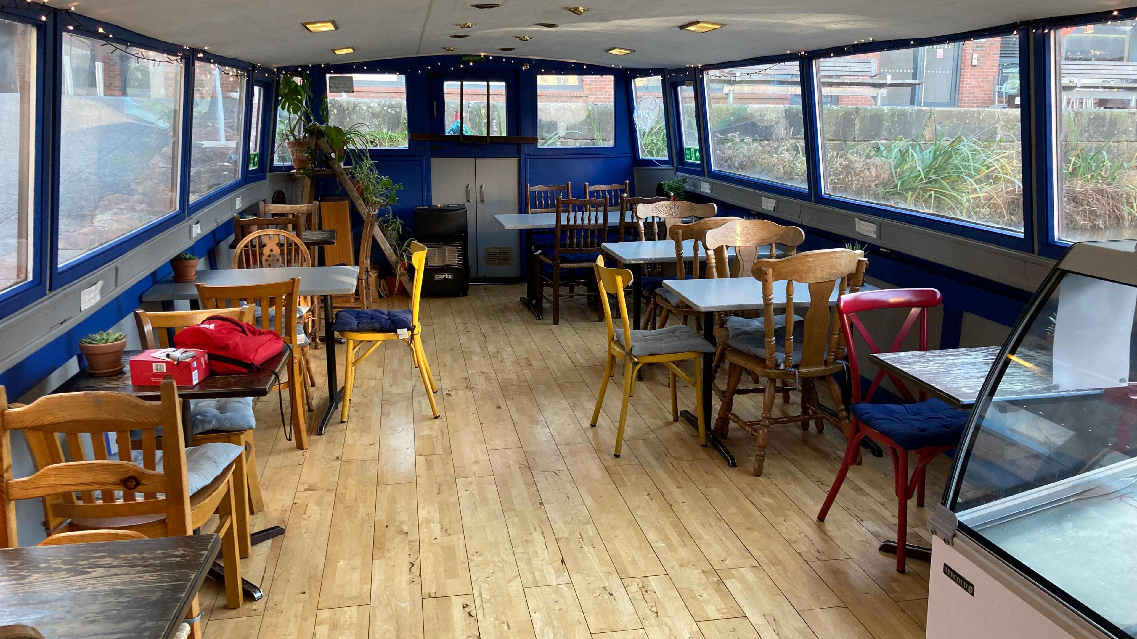 The interior of the narrowboat, which has large windows and blue walls. It has mostly wooden chairs and tables, fairy lights and some plants. 