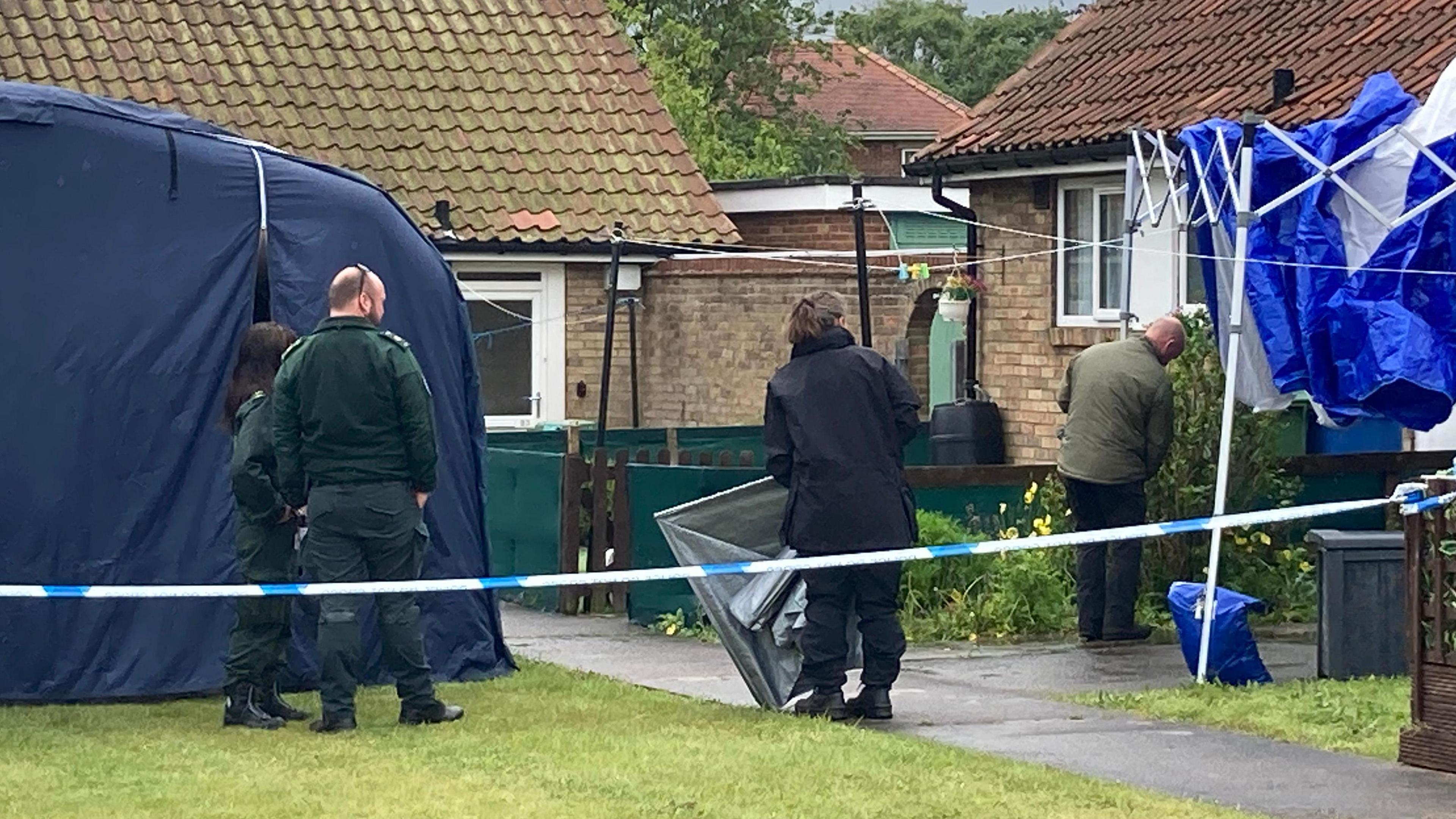 Emergency service workers stand on the grass beside two yellow brick bungalows. There are two officers to the left in dark green trousers and jumpers, and a third to their right in a black coat and trousers, all with their backs to the camera. A fourth person stands on the path to a house in a khaki green jacket. There is a navy tent to the left and a blue and white gazebo to the right, with blue police tape running across the front of the area.