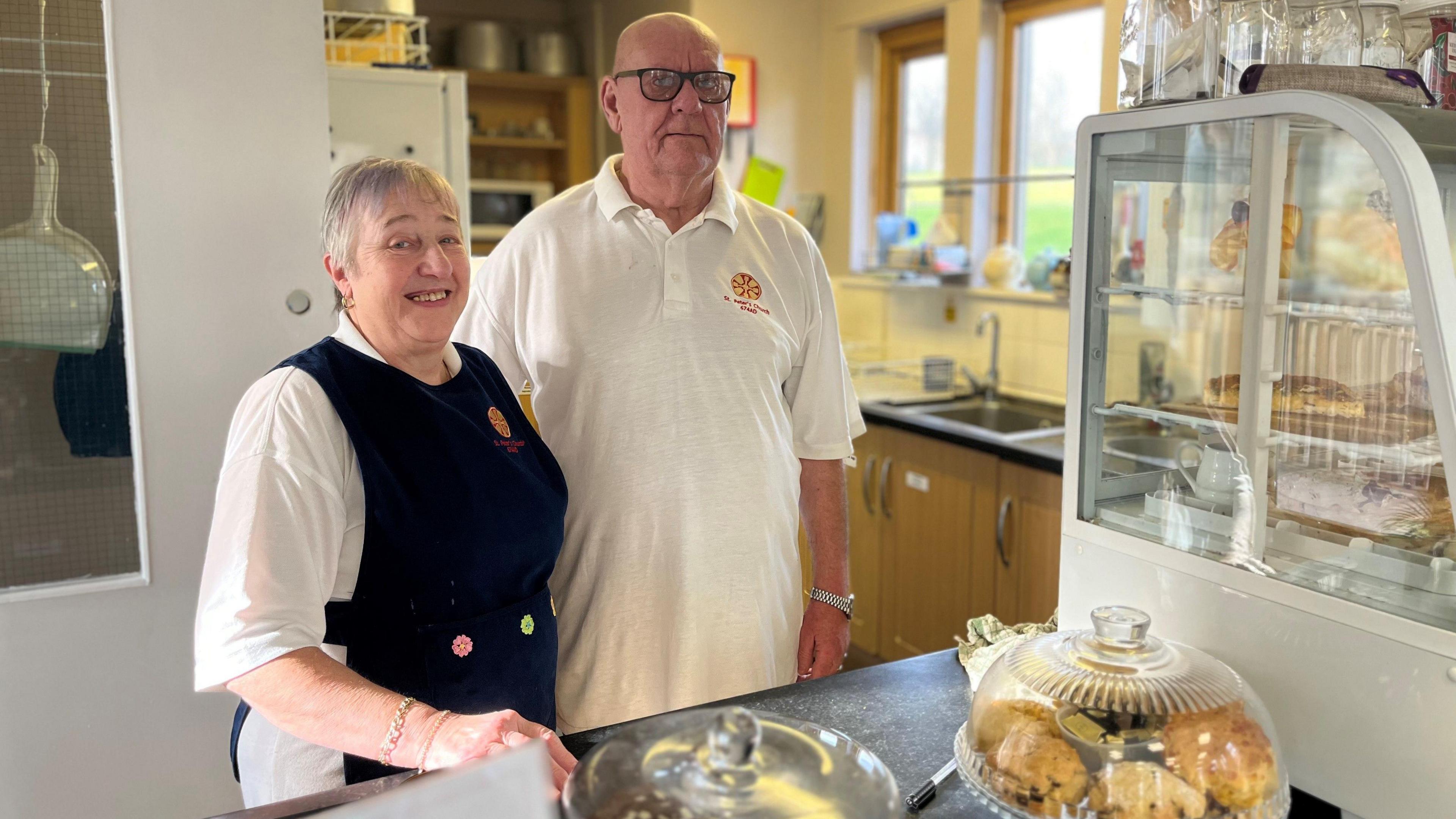 Anne Kemp smiling at the camera next to her husband Derek Kemp behind the counter at Bede's Bakehouse. They are wearing white t-shirts with the red and yellow logo of St Peter's Church. Anne has short grey hair and is wearing a dark blue apron. Derek has black-rimmed glasses. There is a range of cakes and bakes in front of them.