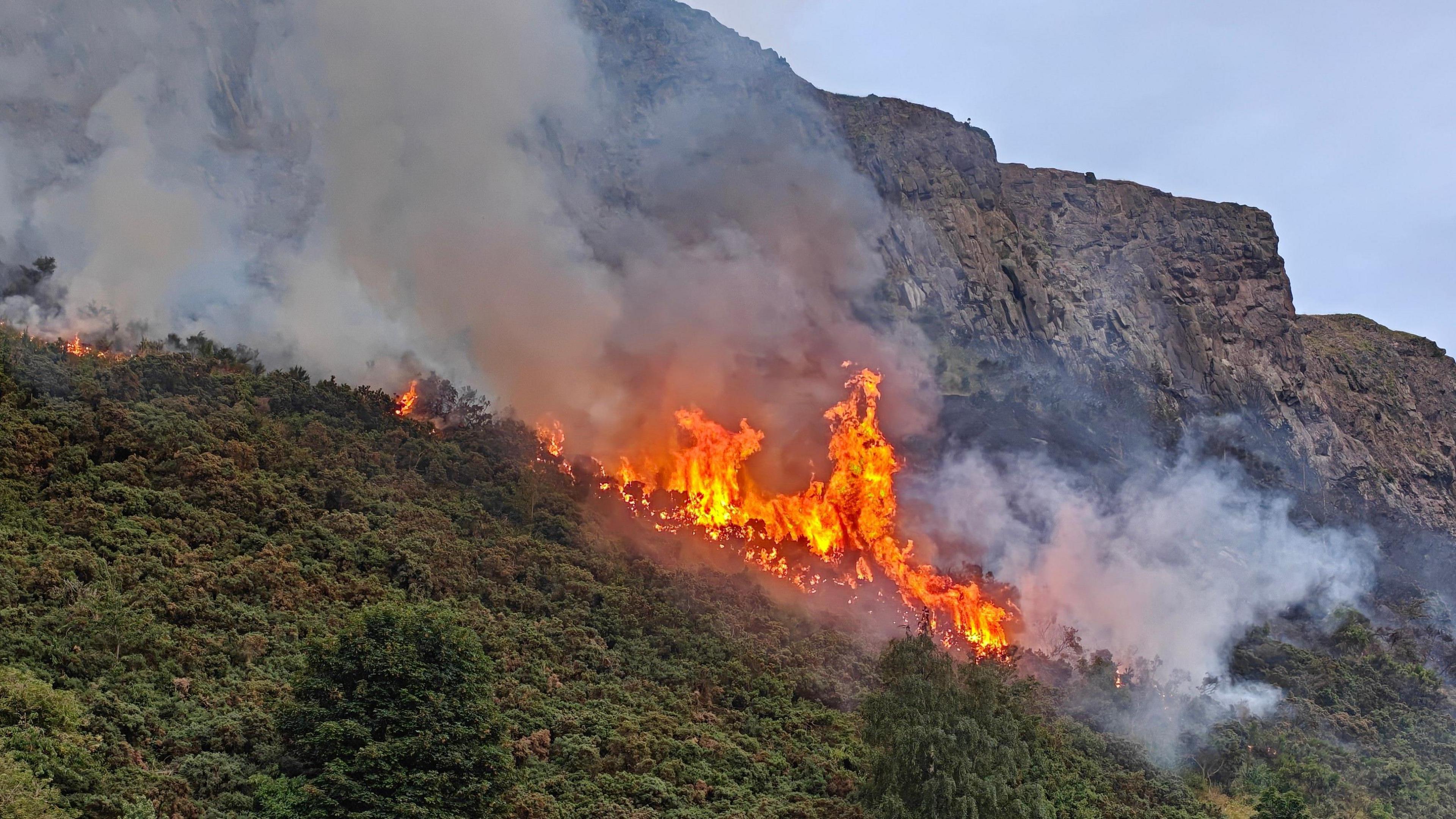 Burning vegetation on the side of Arthur's Seat. There are orange flames and plumes of smoke.