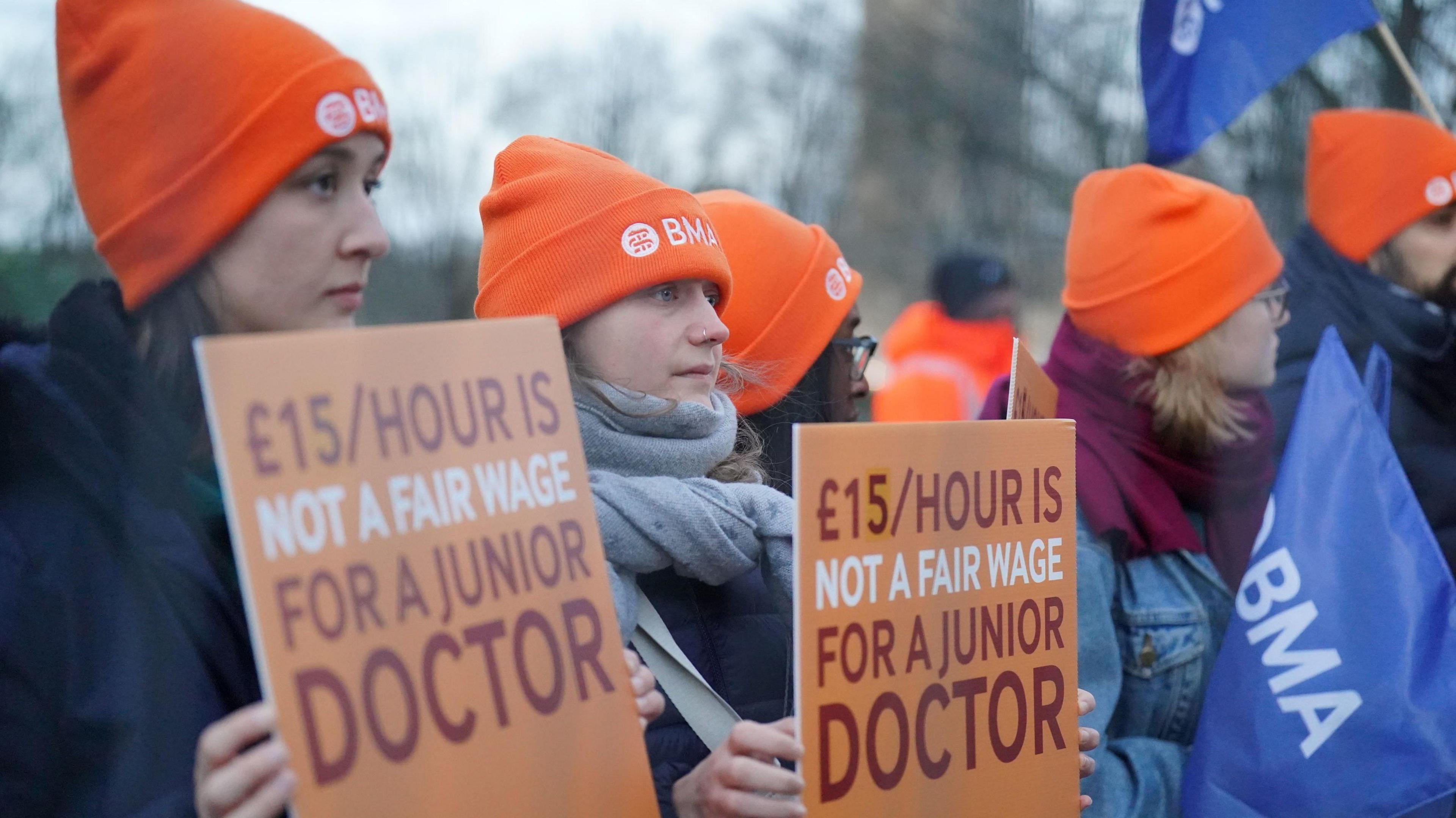 Women in woollen hats hold signs up at a protest reading "£15/hour is not a fair wage for a junior doctor"