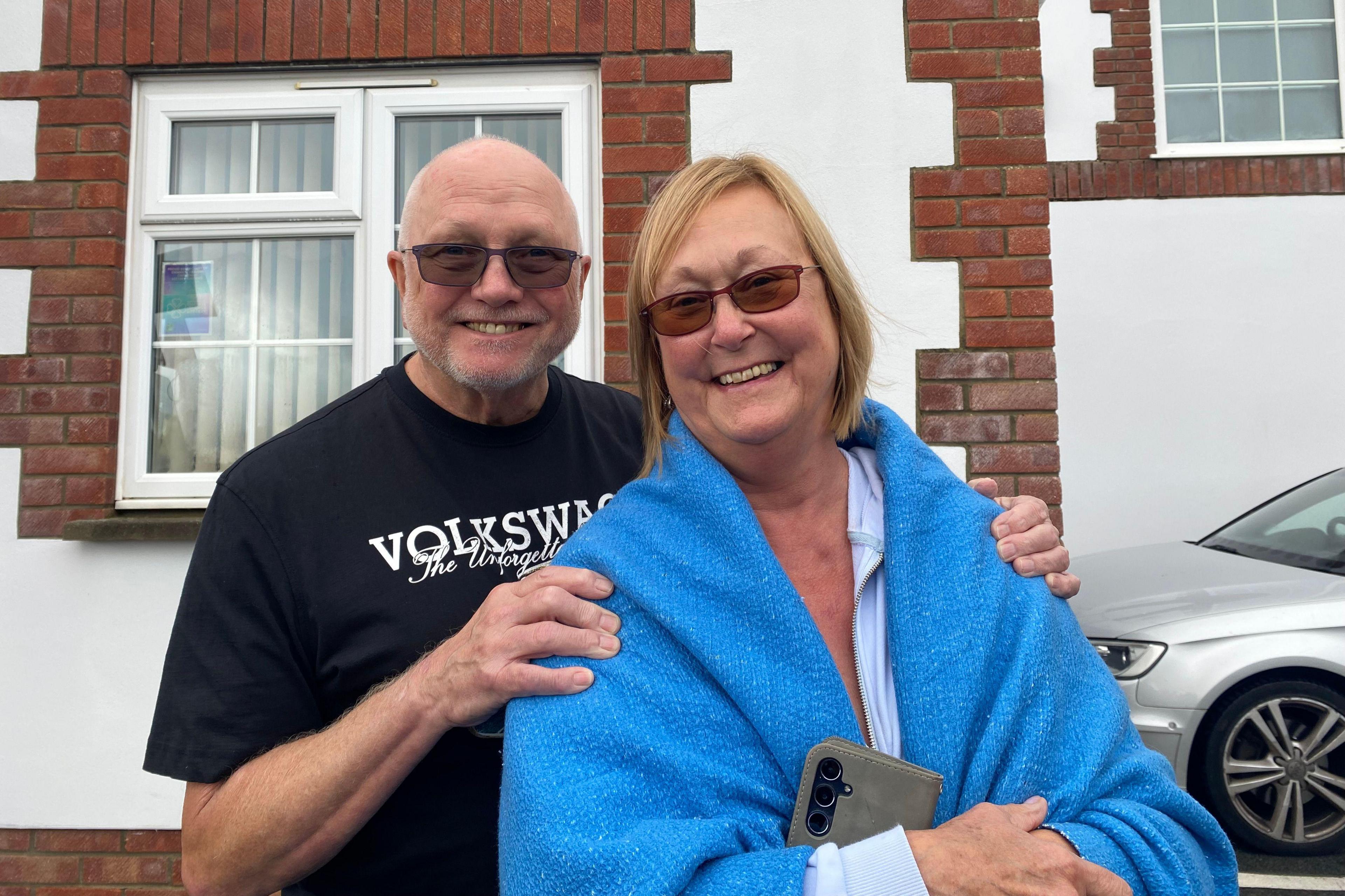 A bald man in sunglasses and a smiling woman with a blonde bob haircut and a blue blanket wrapped around her shoulders hold each other. They stand in front of a white building with brickwork around the windows.