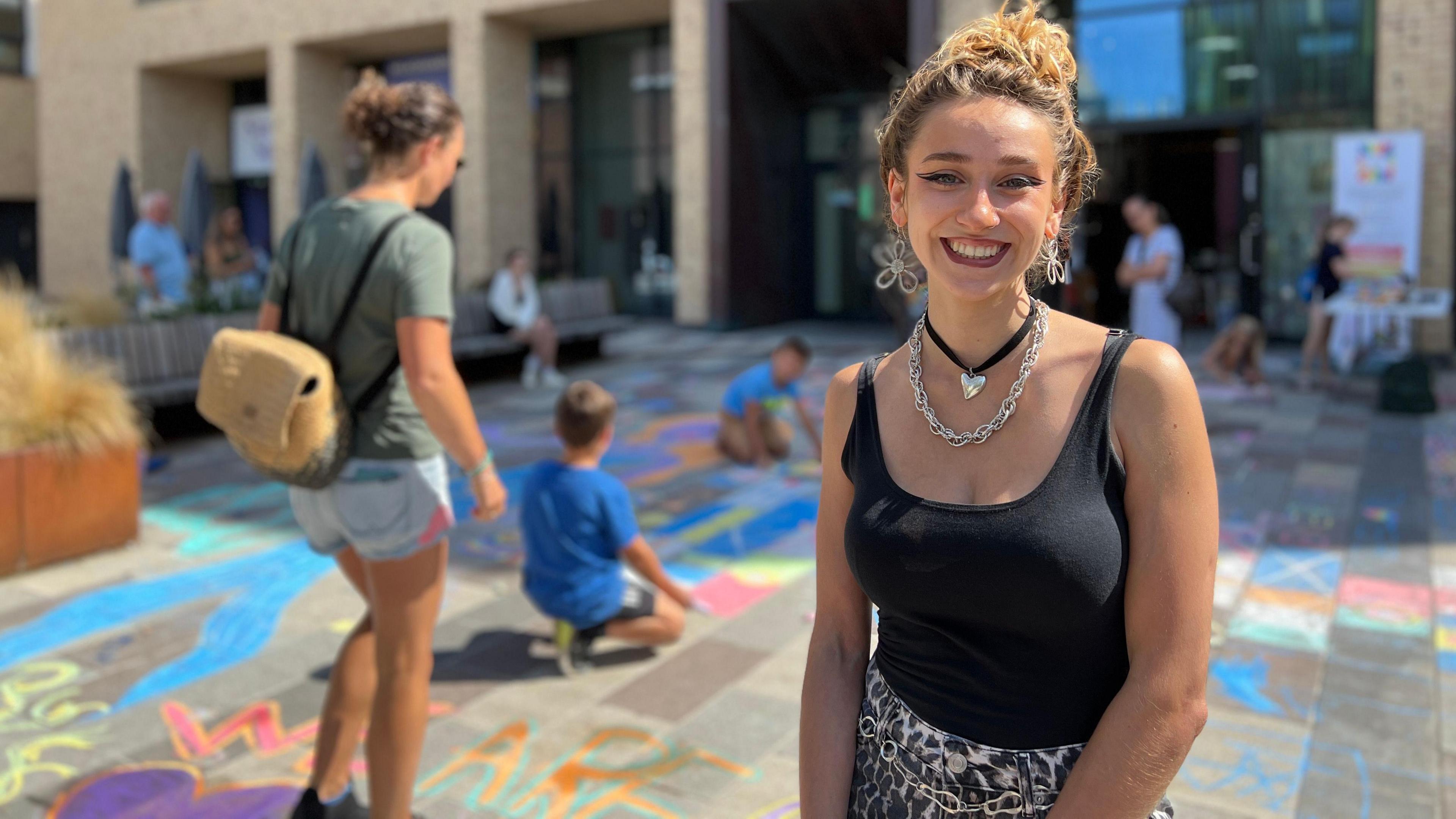 Children and adults participate in a lively outdoor community event in front of a modern building with large windows and balconies. The ground is covered in colorful chalk drawings, with children sitting and drawing while adults stand or walk nearby