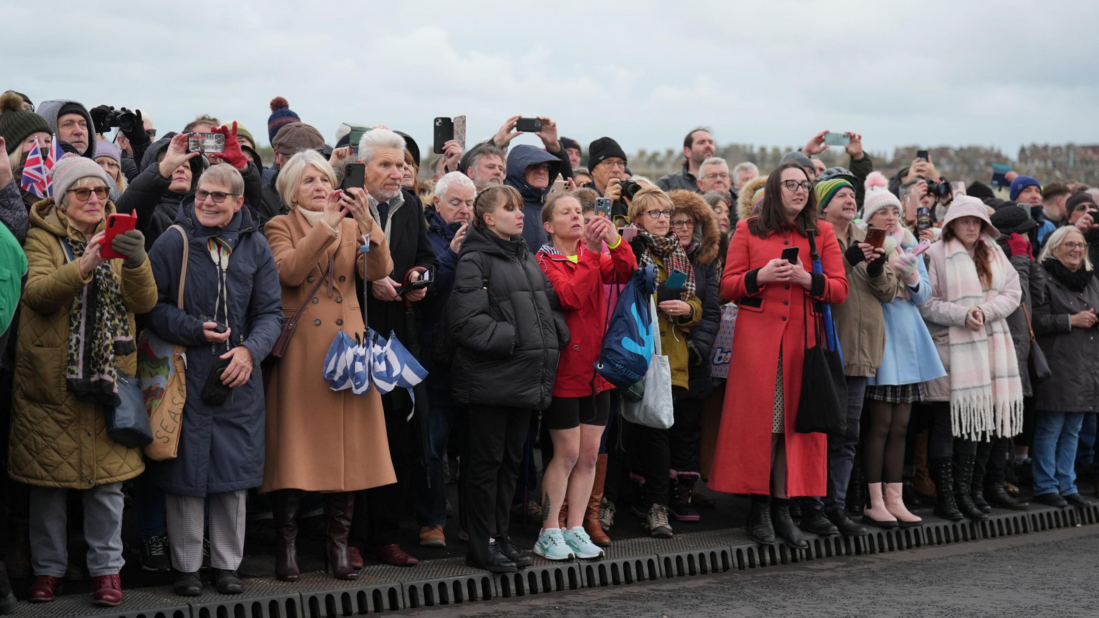 Members of the public wait for the arrival of the Princess Royal to officially open the Gull Wing Bridge in Lowestoft, Suffolk. They are standing on a pavement on the edge of the bridge and many of them are taking pictures of the princess on their phones, while others are applauding