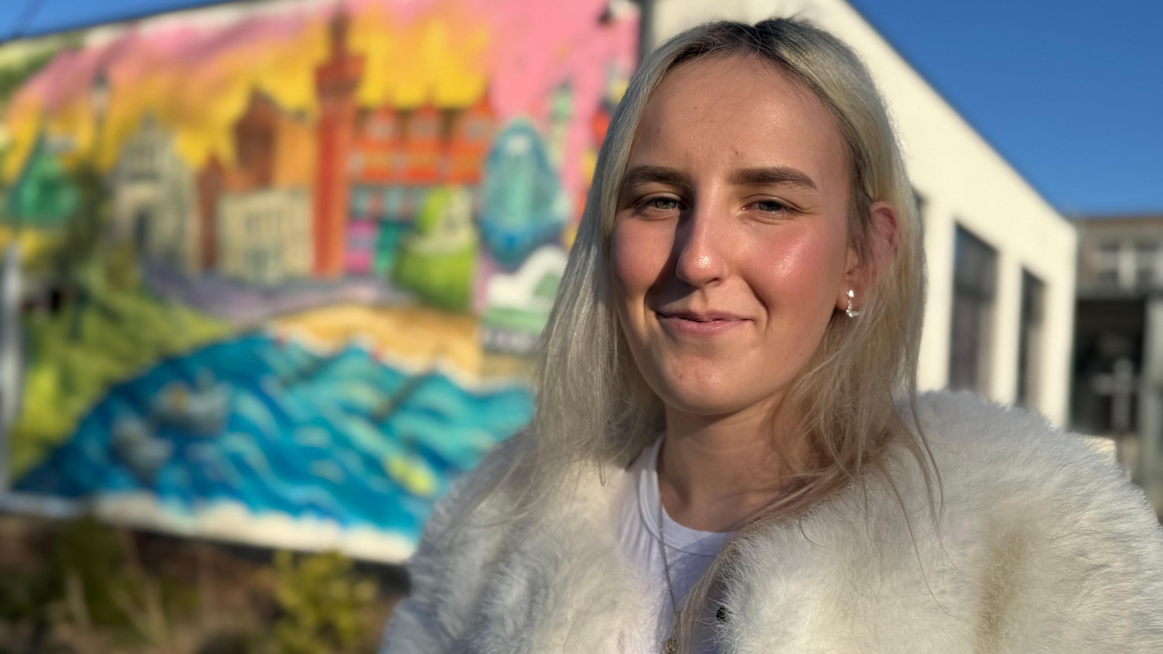 A 19-year-old girl standing in front of a school building with a mural on the side. She has blonde hair and is wearing a furry white coat with white t-shirt and is smiling to the camera.