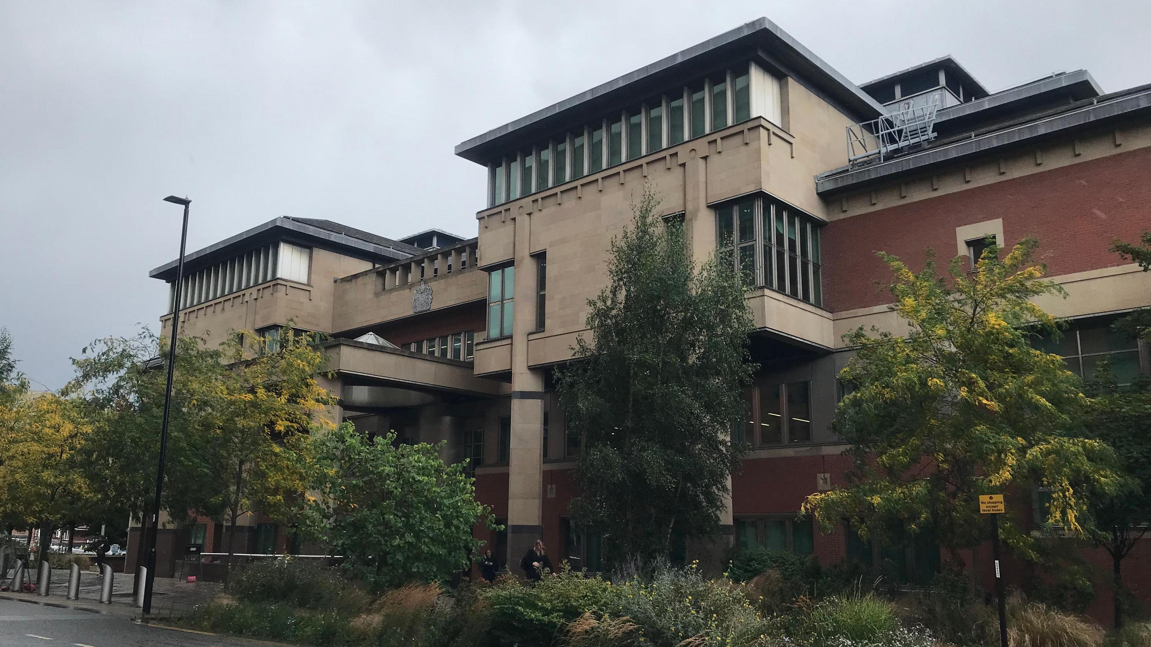 A modern multi-storey building with a mix of beige and red-brick exterior walls. Its architectural design includes large windows and several protruding sections. In front of the building, there are trees and shrubs lining the sidewalk. The sky is overcast, hinting at cloudy or rainy weather, and the street surface appears wet. Street lamps and signage are also visible along the road.