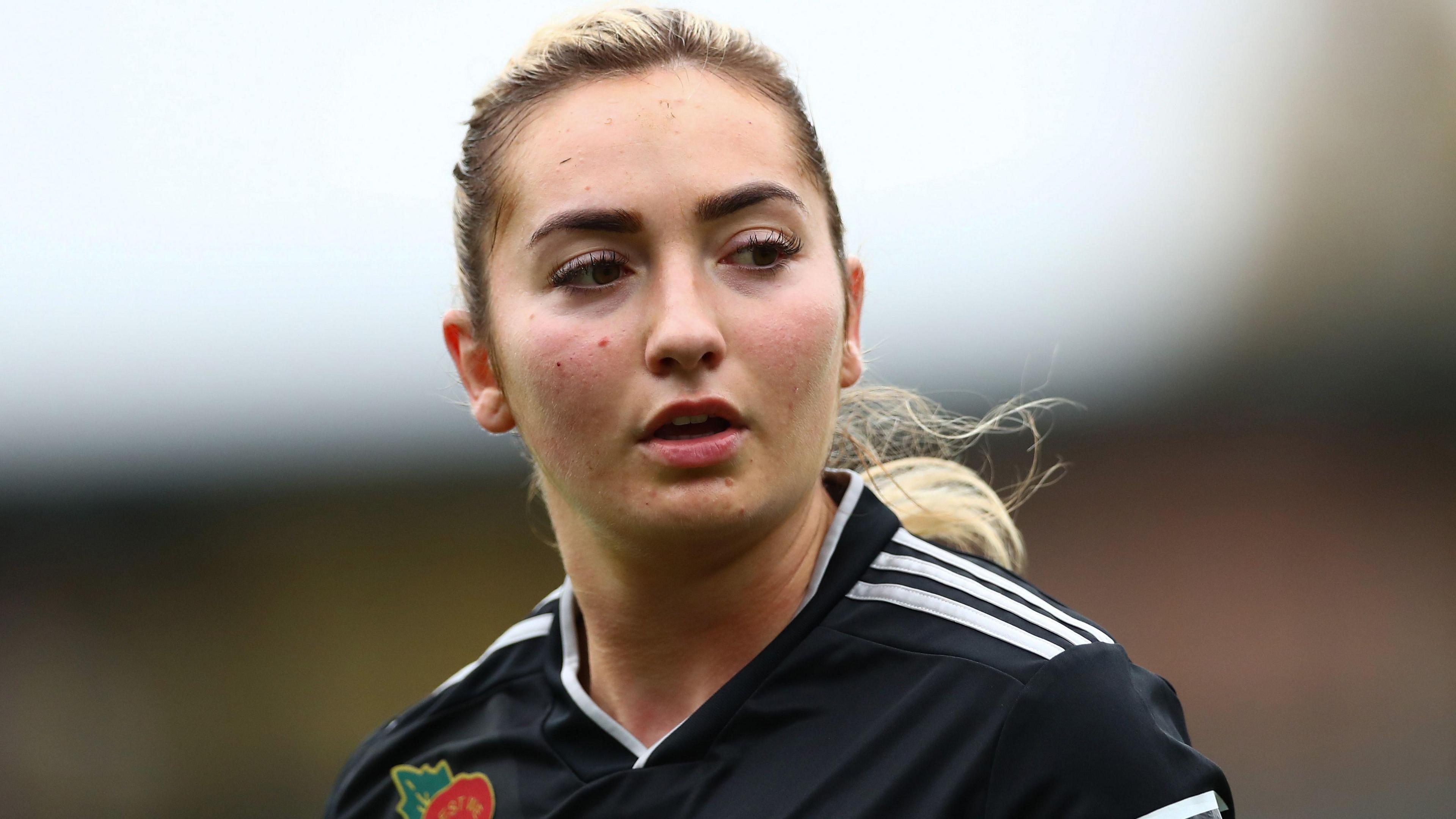 Maddy Cusack playing for Sheffield United in 2021. Maddy has her long blonde hair tied back in a ponytail and wears a black football shirt with three white stripes on the shoulders. She's looking over her left shoulder with a serious expression. 