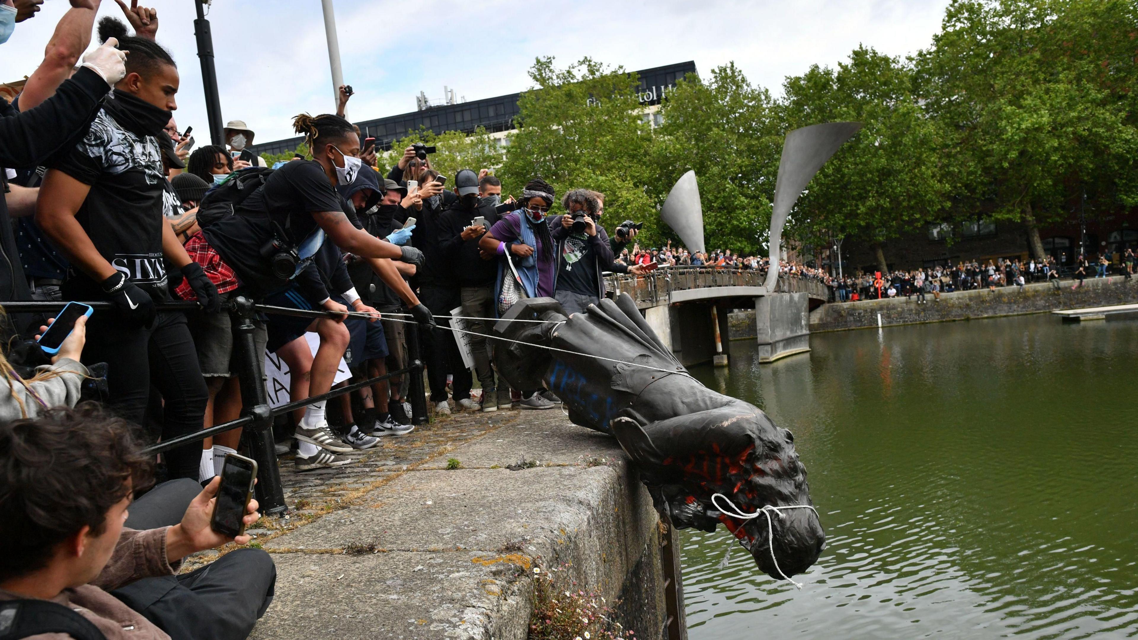 A metal statue of a man in a long coat with long hair being lowered on ropes over the side of a harbour, towards the water. There are hundreds of people visible watching.