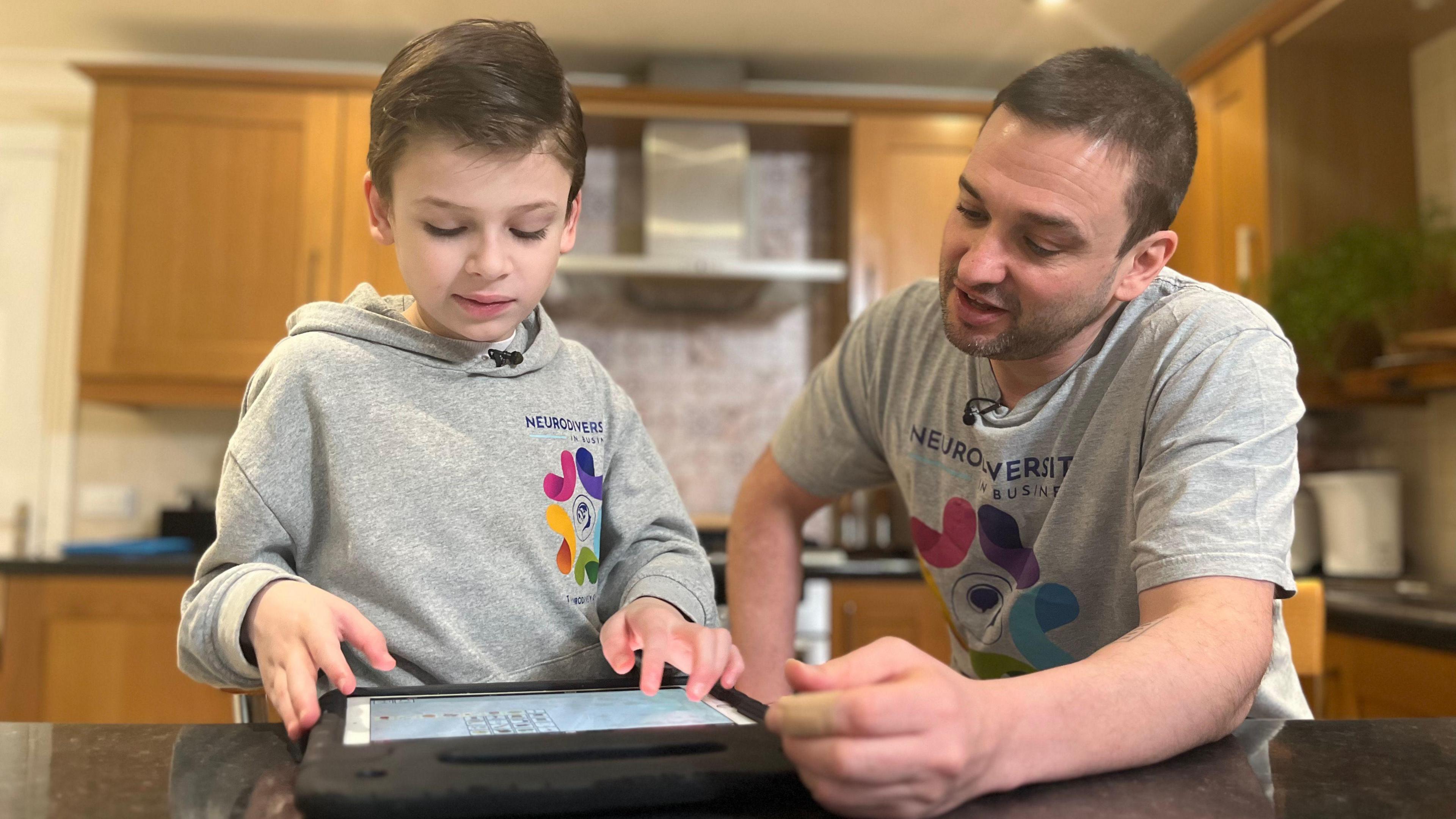 Joshie wearing a grey sweatshirt looking at the ipad next to Dan wearing a grey T-shirt .