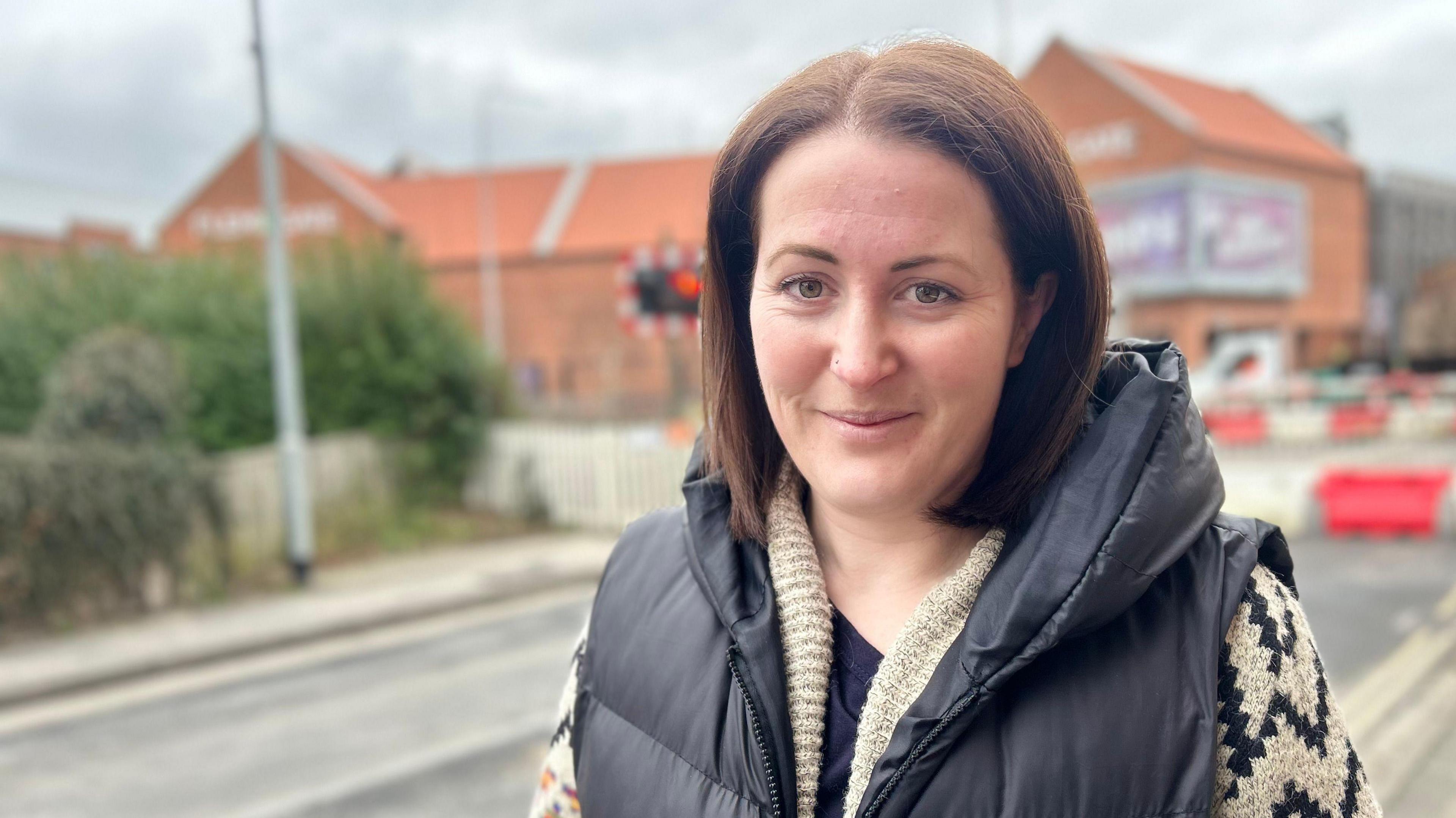 Helena Wilson is standing on a pavement near a road closure caused by roadworks. A railway level crossing and the buildings of red-brick shopping centre can be seen. Helena has shoulder-length brown hair and wears a dark gilet and cream cardigan. 