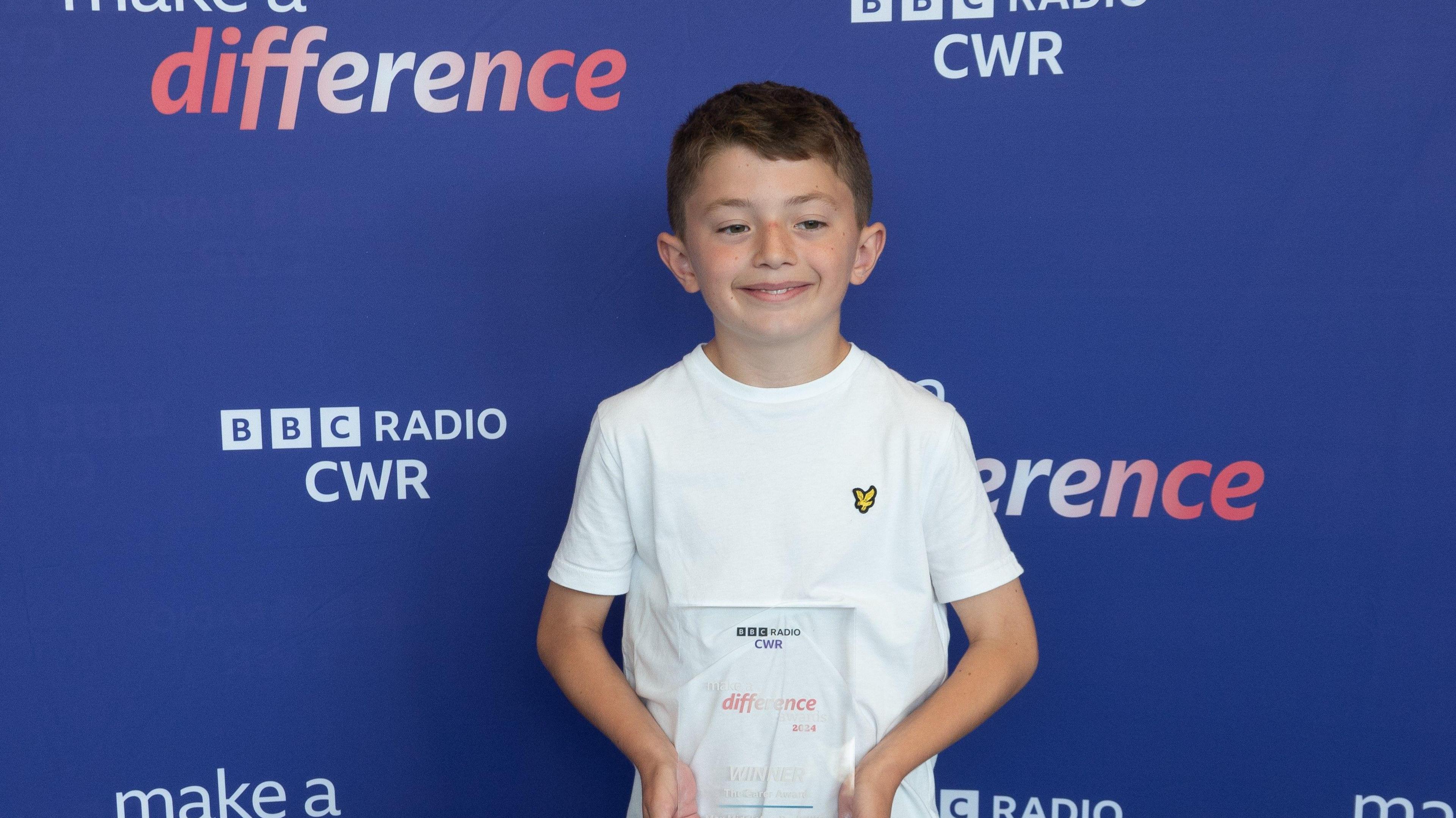 A young boy stands in front of a blue background holding a make a difference award