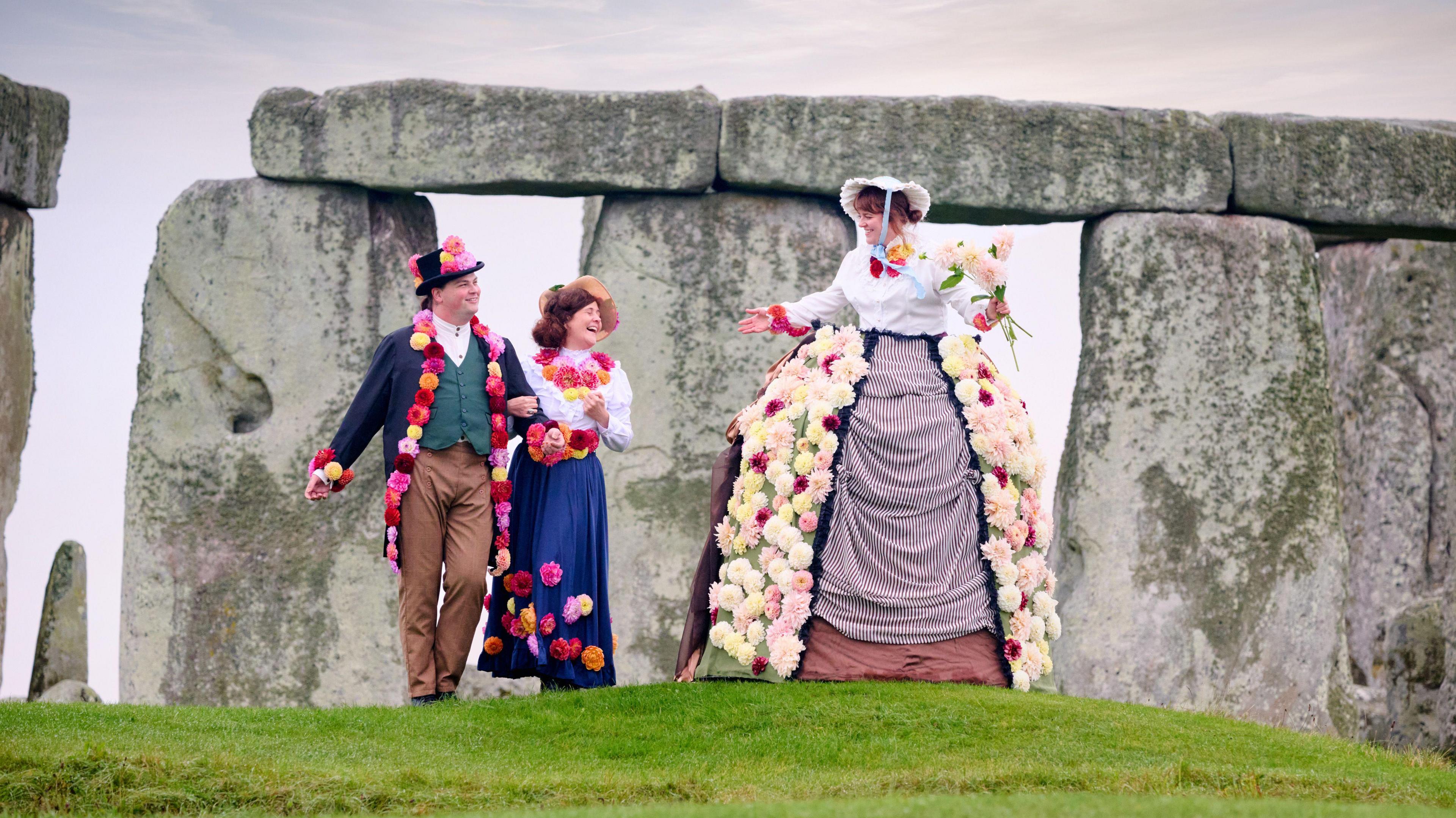 A tall woman wearing a large skirt which is decorated with dahlias. She is wearing Victorian style clothing and a bonnet, and is walking alongside a couple with her arms outstretched and carrying a bouquet of dahlias. The couple are linking arms, also wearing Victorian clothing adorned with colourful dahlias.