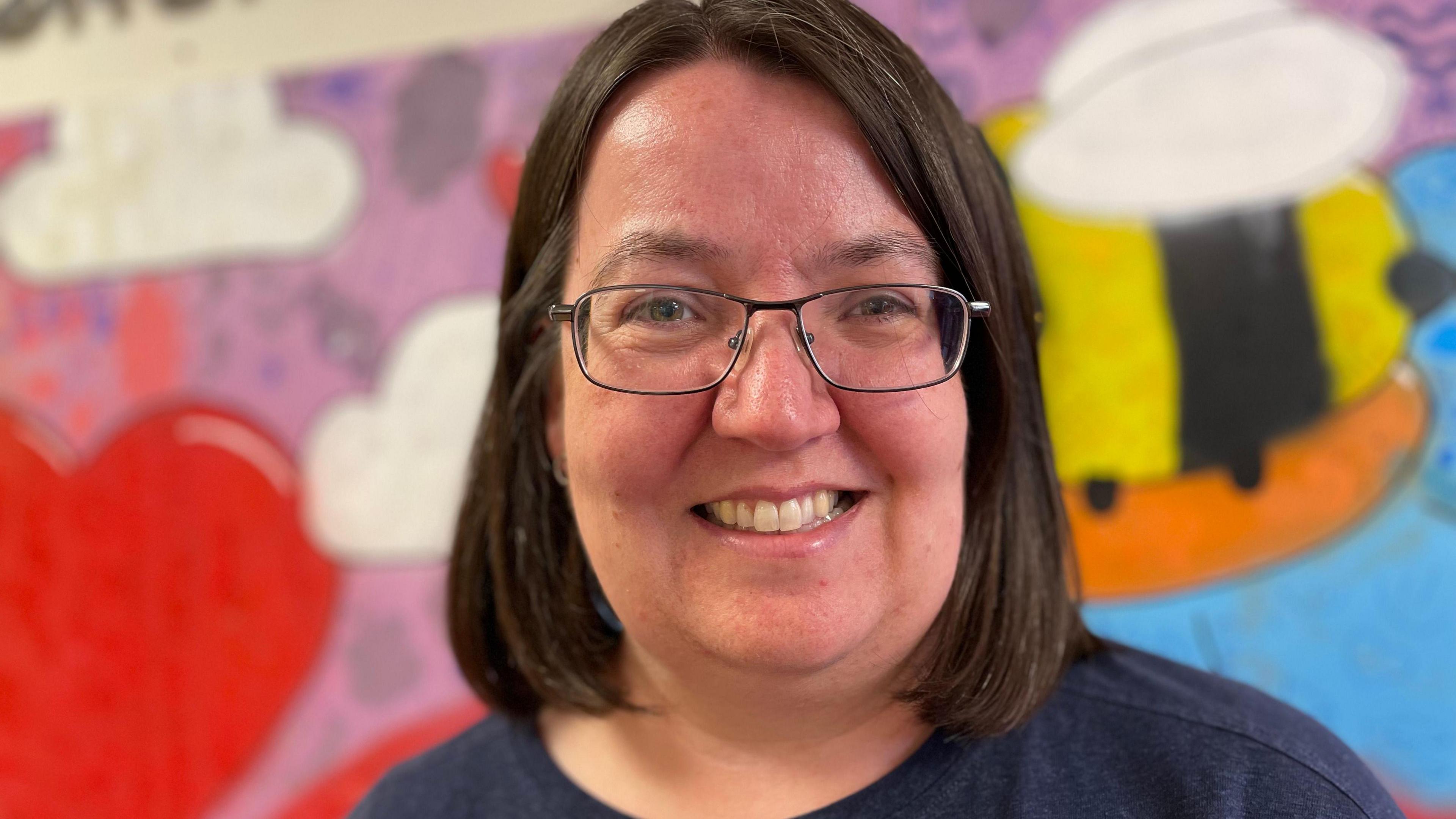 A close-up of a woman with her hair in a brown bob. She is smiling and wearing glasses and a navy blue top. She is standing in front of a colourful mural, which includes red love hearts and a black and yellow bee. 