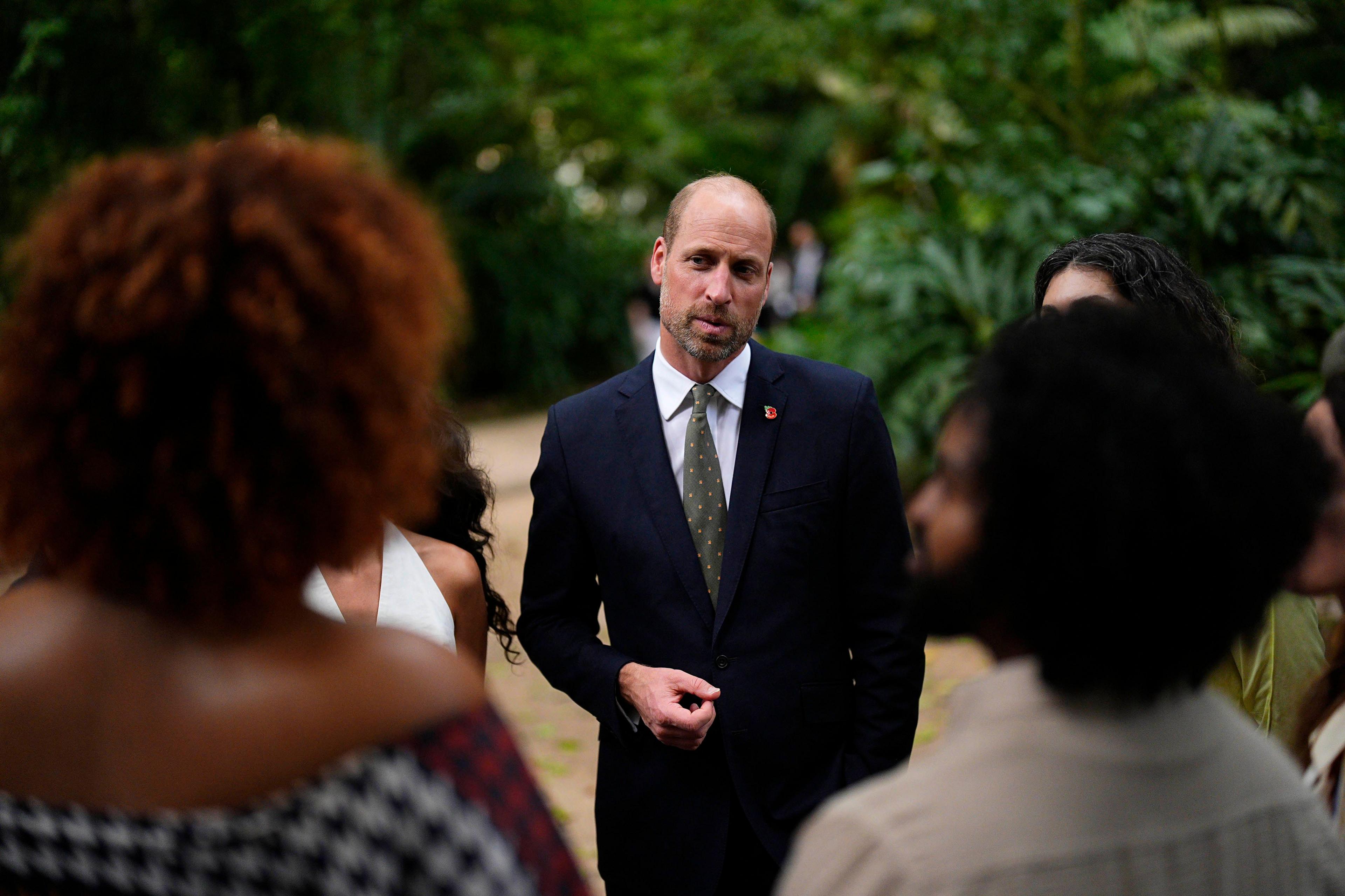 William, dressed in a dark suit, white shirt and tie, is facing camera as he stands on a path in the jungle looking quizzically at two people on either side of him with their back to the camera.