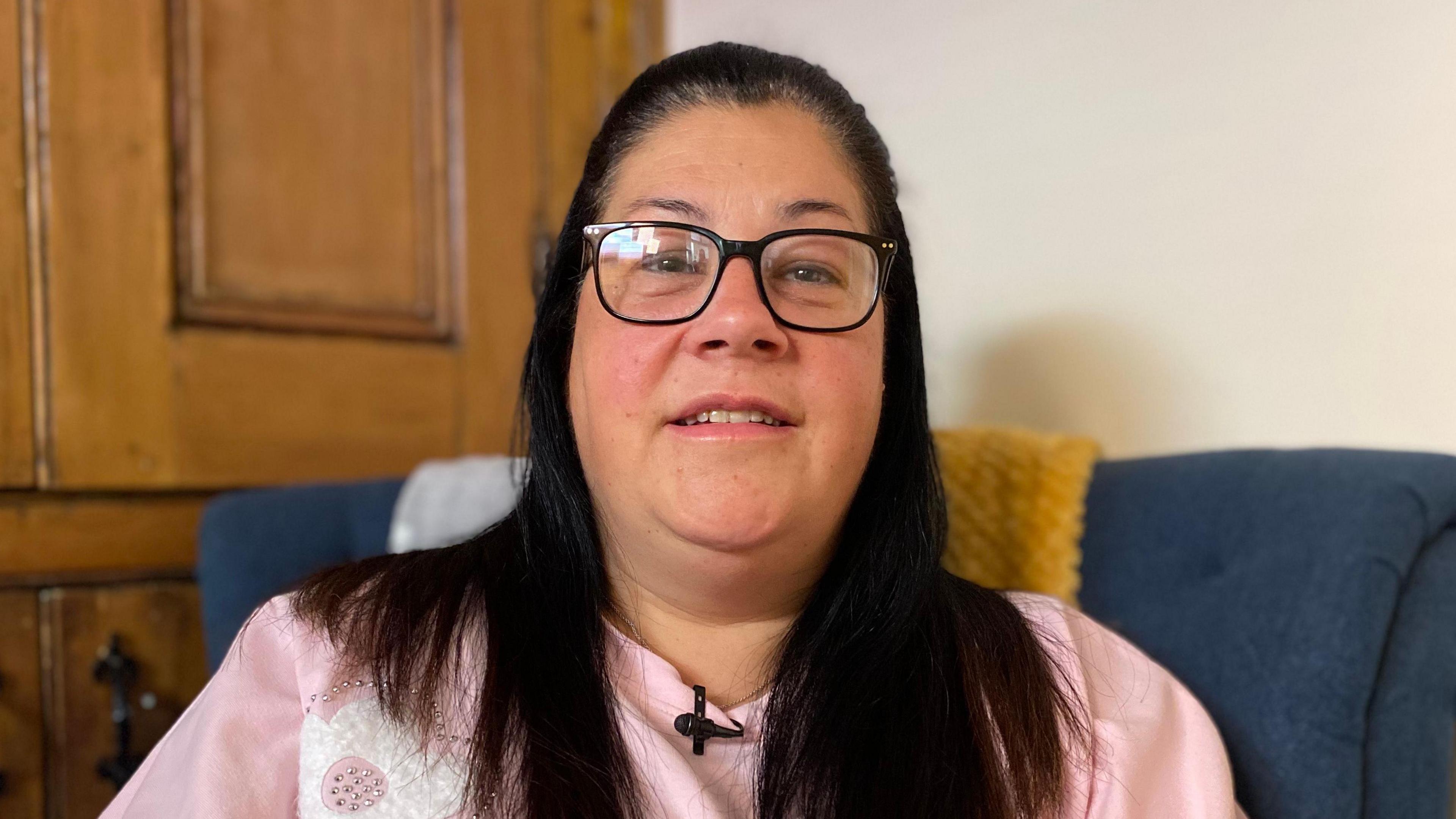 Rachael Wild sitting in a blue chair. She is wearing glasses and a pinky top with white and silver embellishment