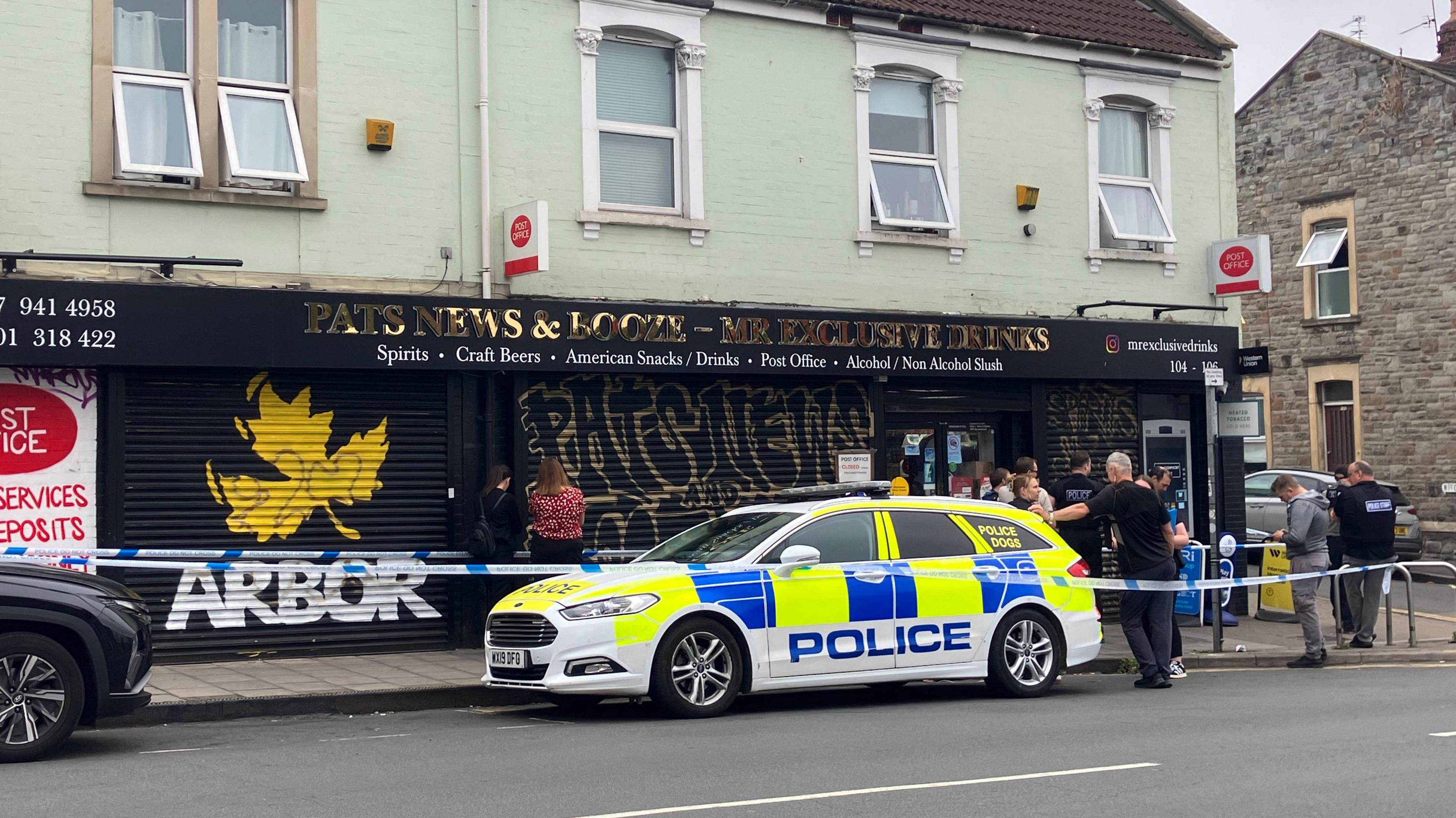 A police car parked outside an off-licence shop. There is blue and white police tape and several officers standing round outside the shop.