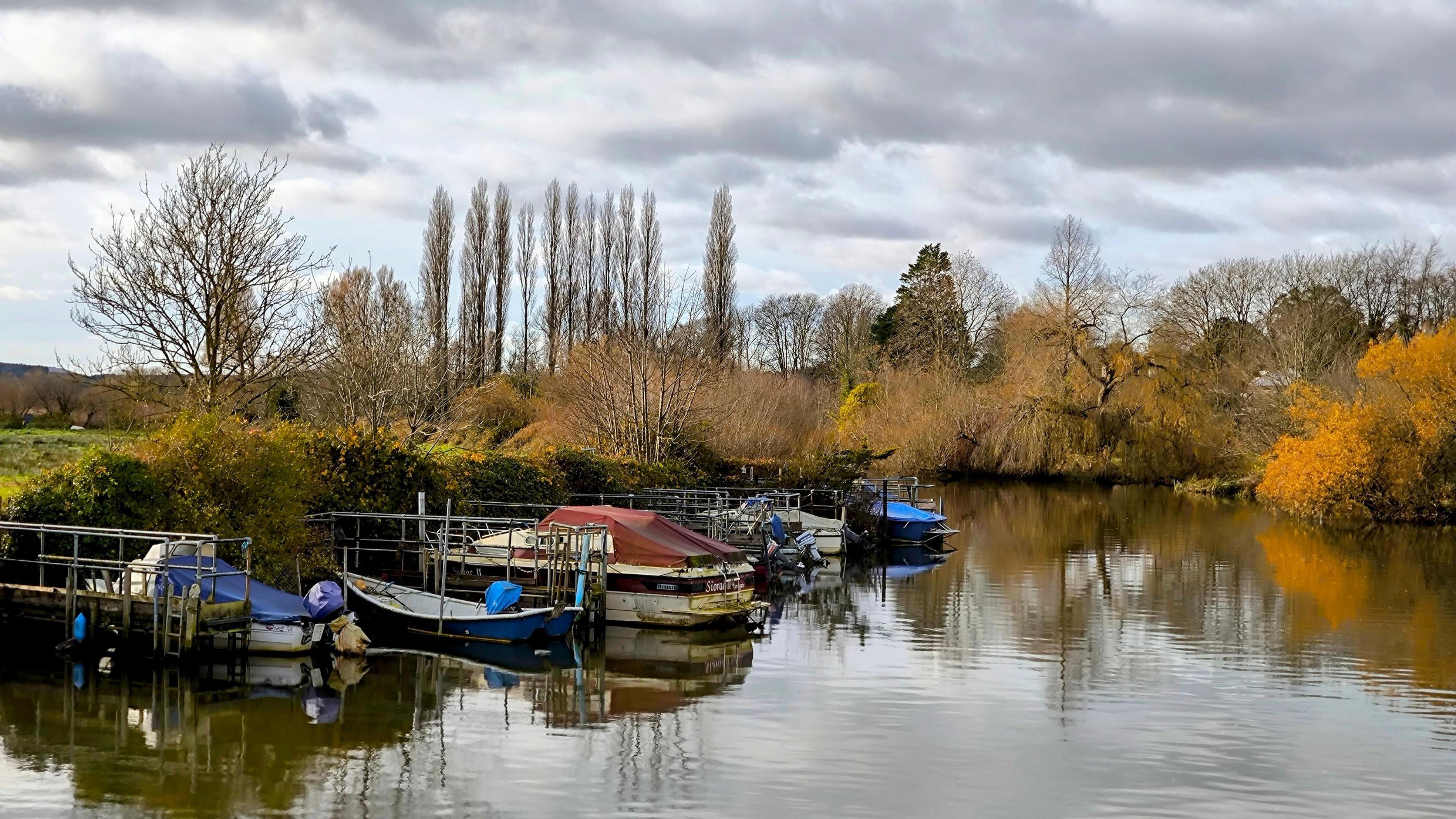 A wintery river scene with boats moored at the bank. The trees lining the river have no leaves and the sky has hazy sunshine with grey clouds.