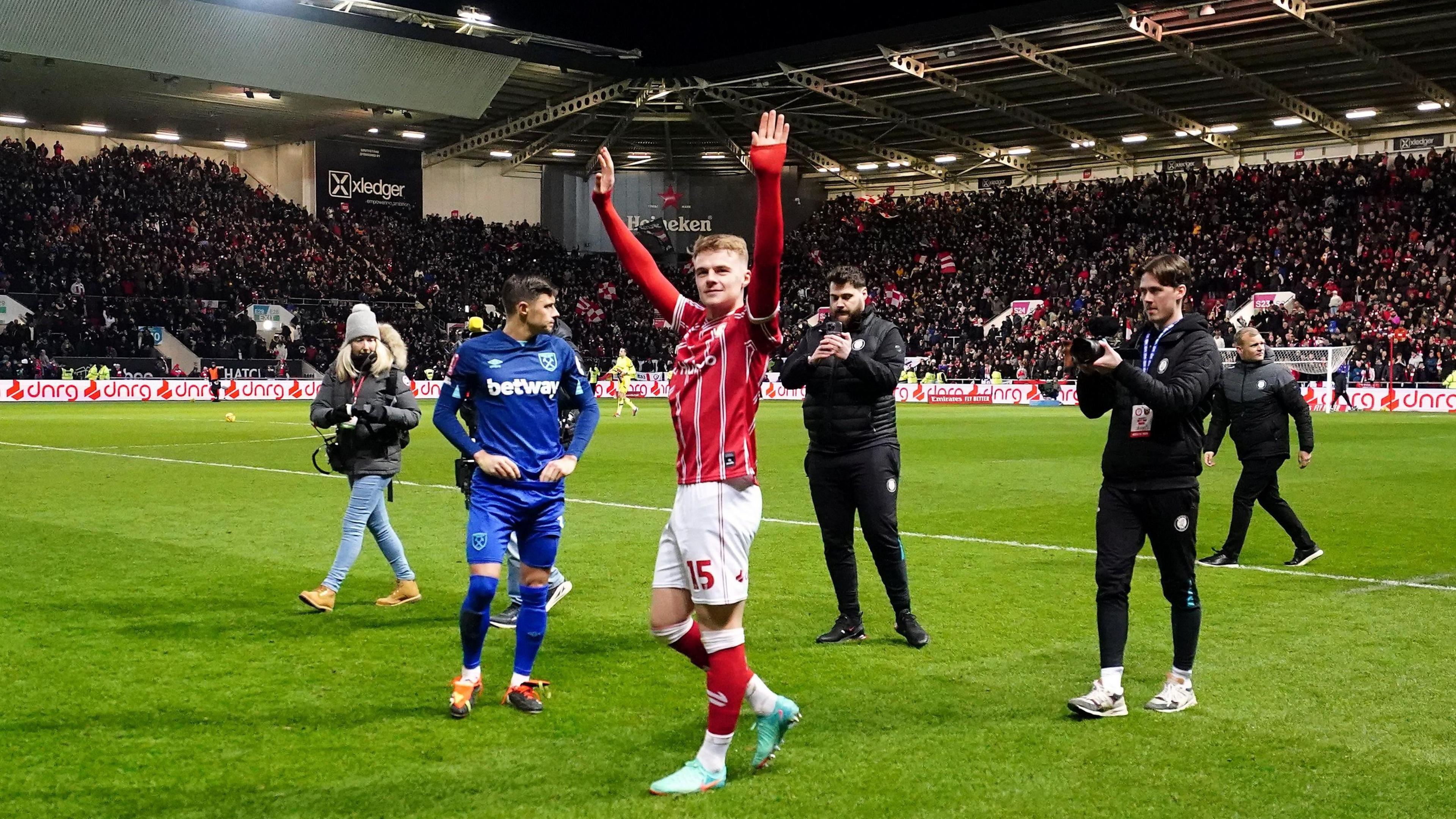 Bristol City player Tommy Conway celebrates with his hands in the air after City beat West Ham in the FA Cup at Ashton Gate. He is being followed by two men who are filming him with their phones and in the background the stands are still full of fans.