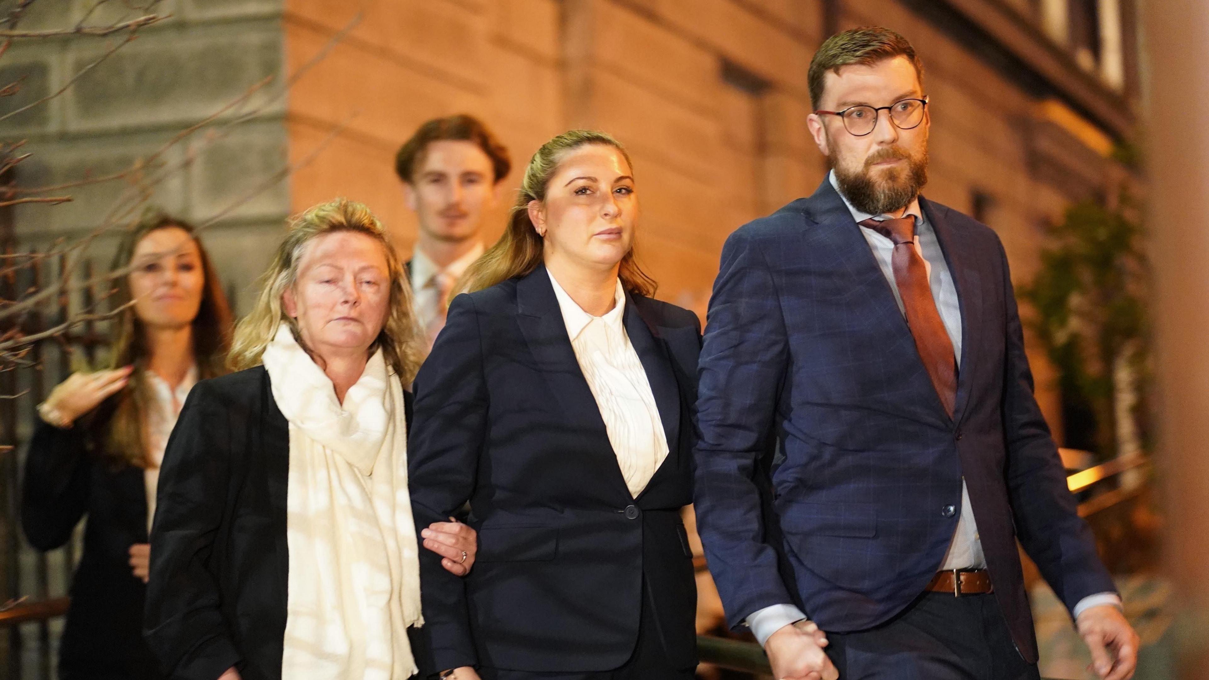 From left to right, there is a woman with blond hair, wearing a black jacket and white scarf, a woman with blond hair, wearing white shirt and black blazer and a man with brown hair and a beard. He is wearing glasses, a navy suit, blue shirt and red tie. There are two people blurred in the background. 