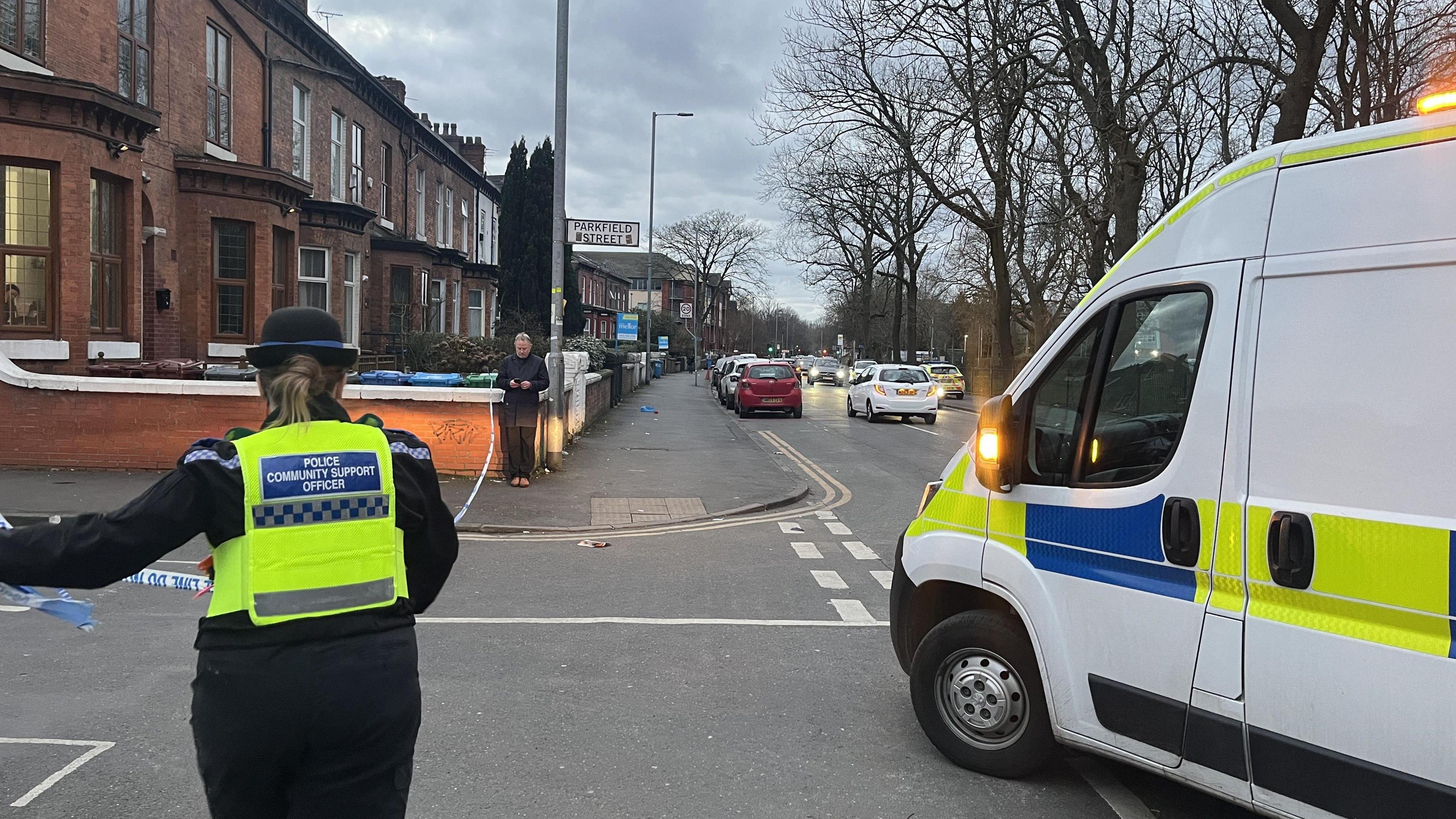 A white police van with yellow and blue markings is guided into a road by a hi-vis wearing Police Community Support Officer.