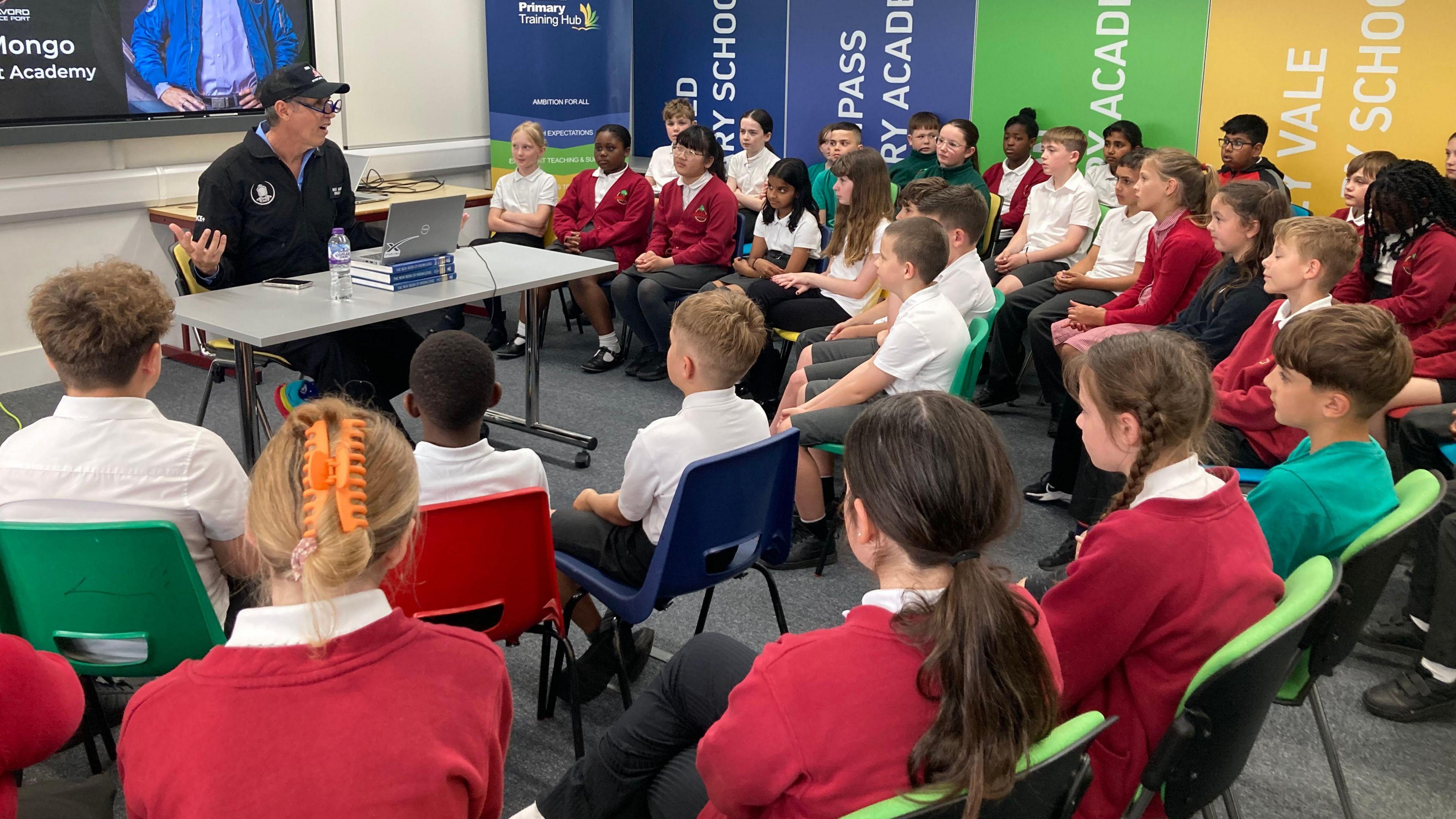 An astronaut teacher in his black space suit speaks to lots of year 5 pupils in their red school uniforms