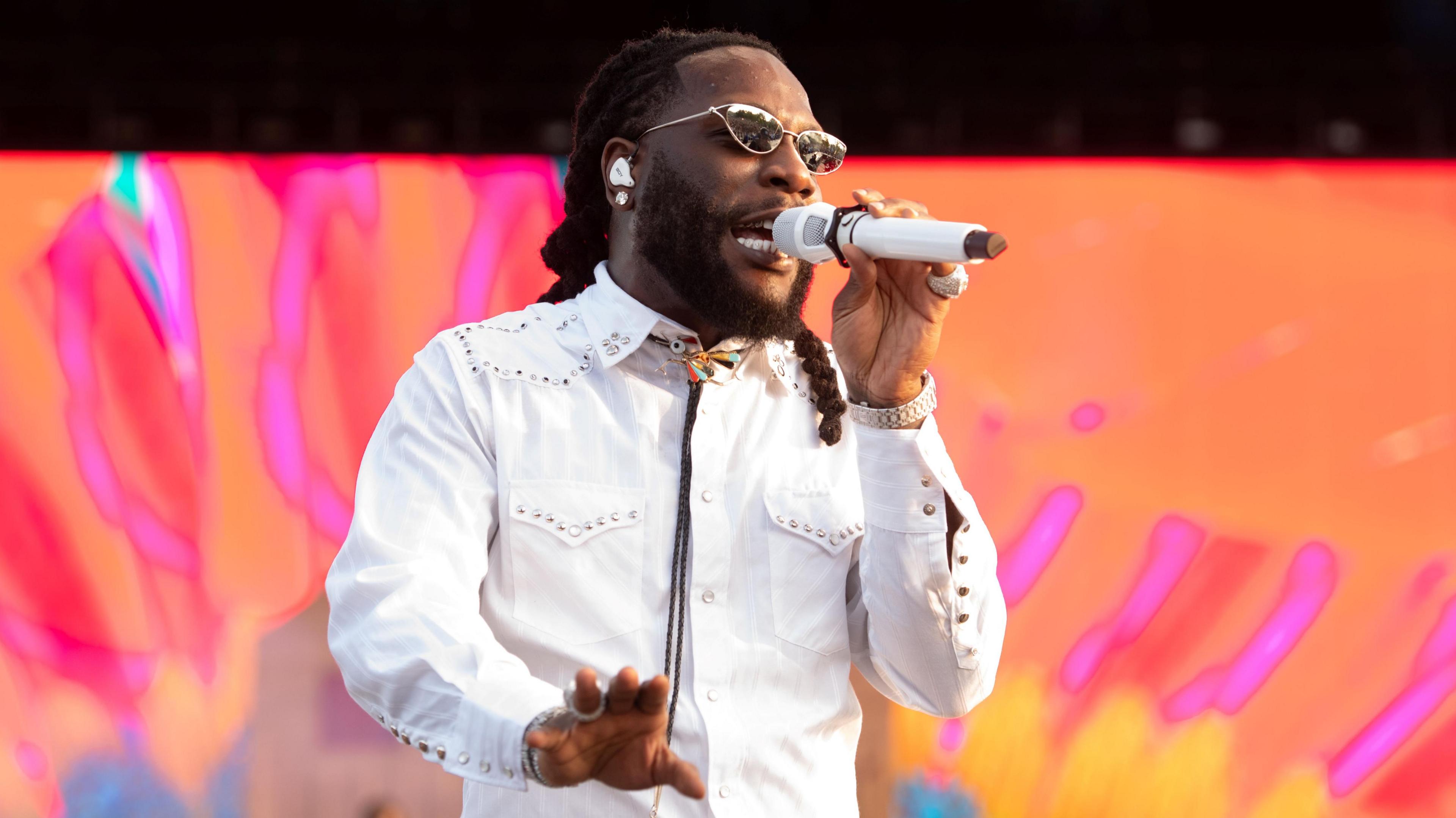 Burna Boy performing at Wireless Festival in July. The singer wears a white Western-style shirt with a black bolo tie. He has braided dark hair and a short beard and is pictured wearing reflective sunglasses. He sings into a mic held in his left hand, his right outstretched towards the crowd. The staging behind him is lit pink and orange.