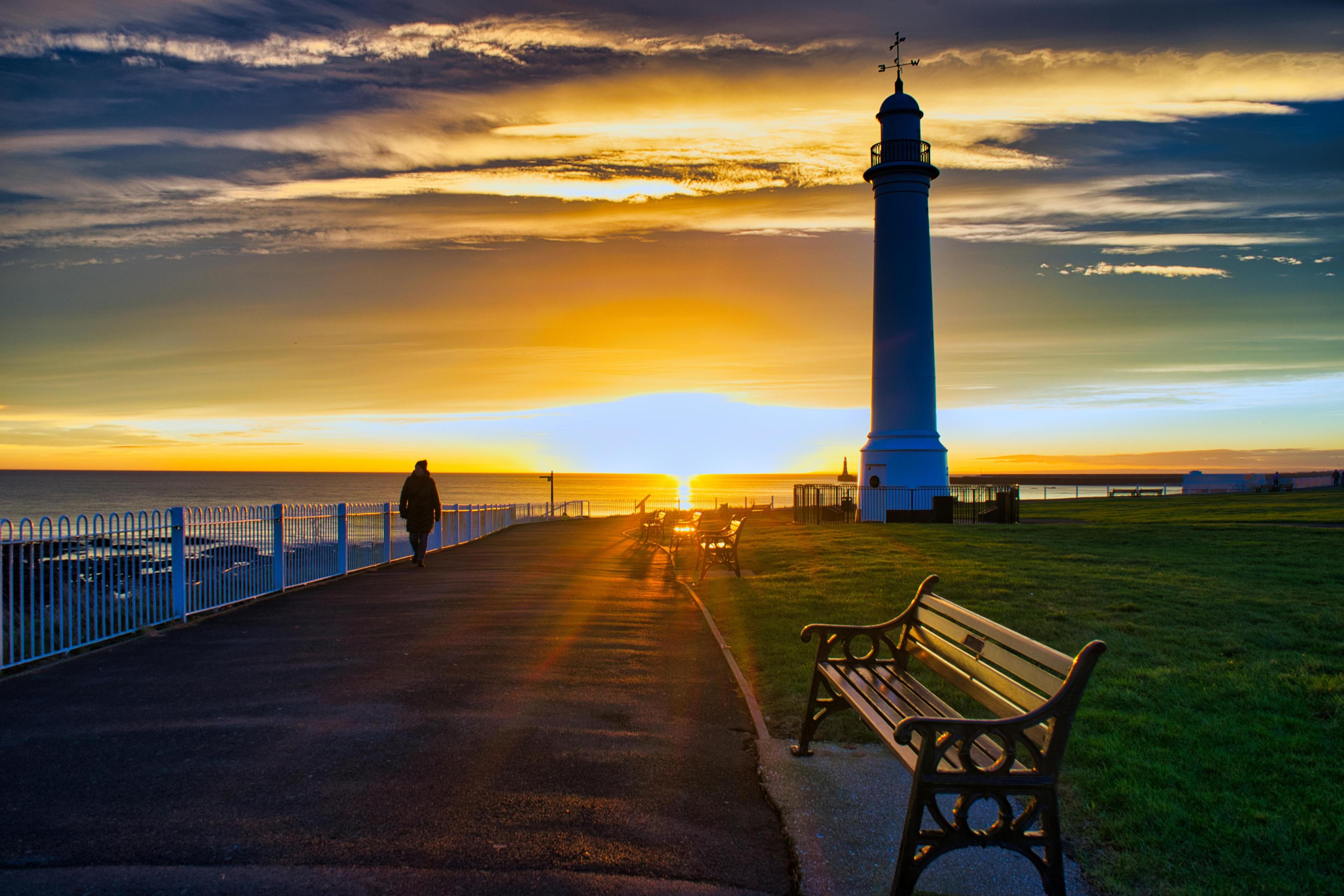 A sunset casts a silhouette of a lighthouse and a person walking along the beach.