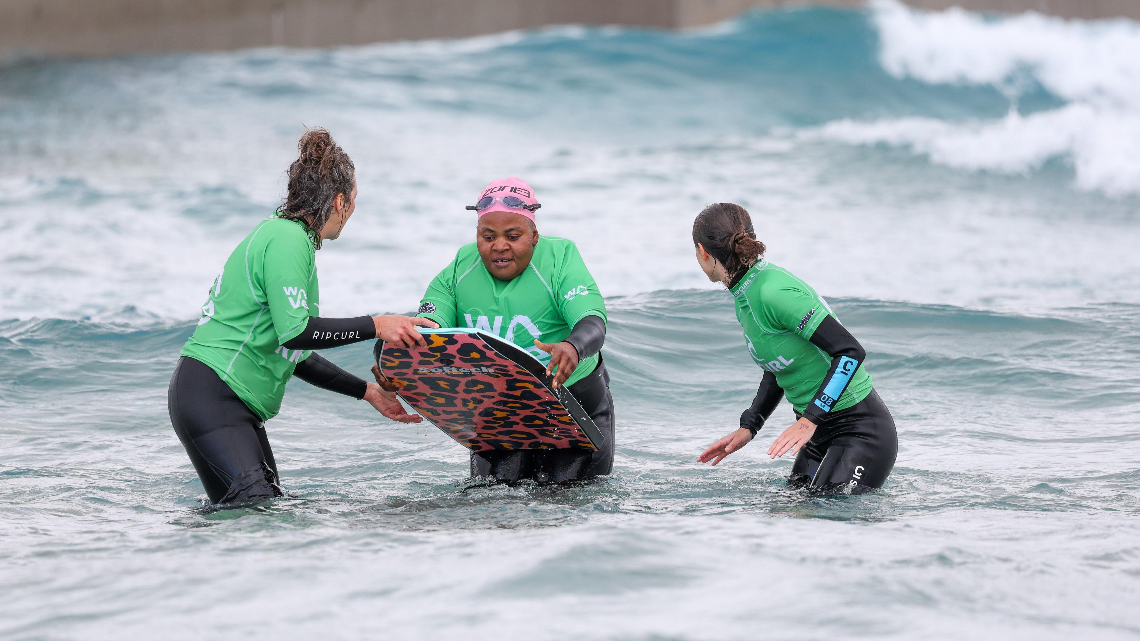 A participant getting ready to catch a wave on her bodyboard, joined on either side by two volunteers who are gearing up to help push her into the wave. The participant is wearing a black wetsuit, green rash vest and pink swimming cap with goggles on her head. Behind them there are two small waves about to break.