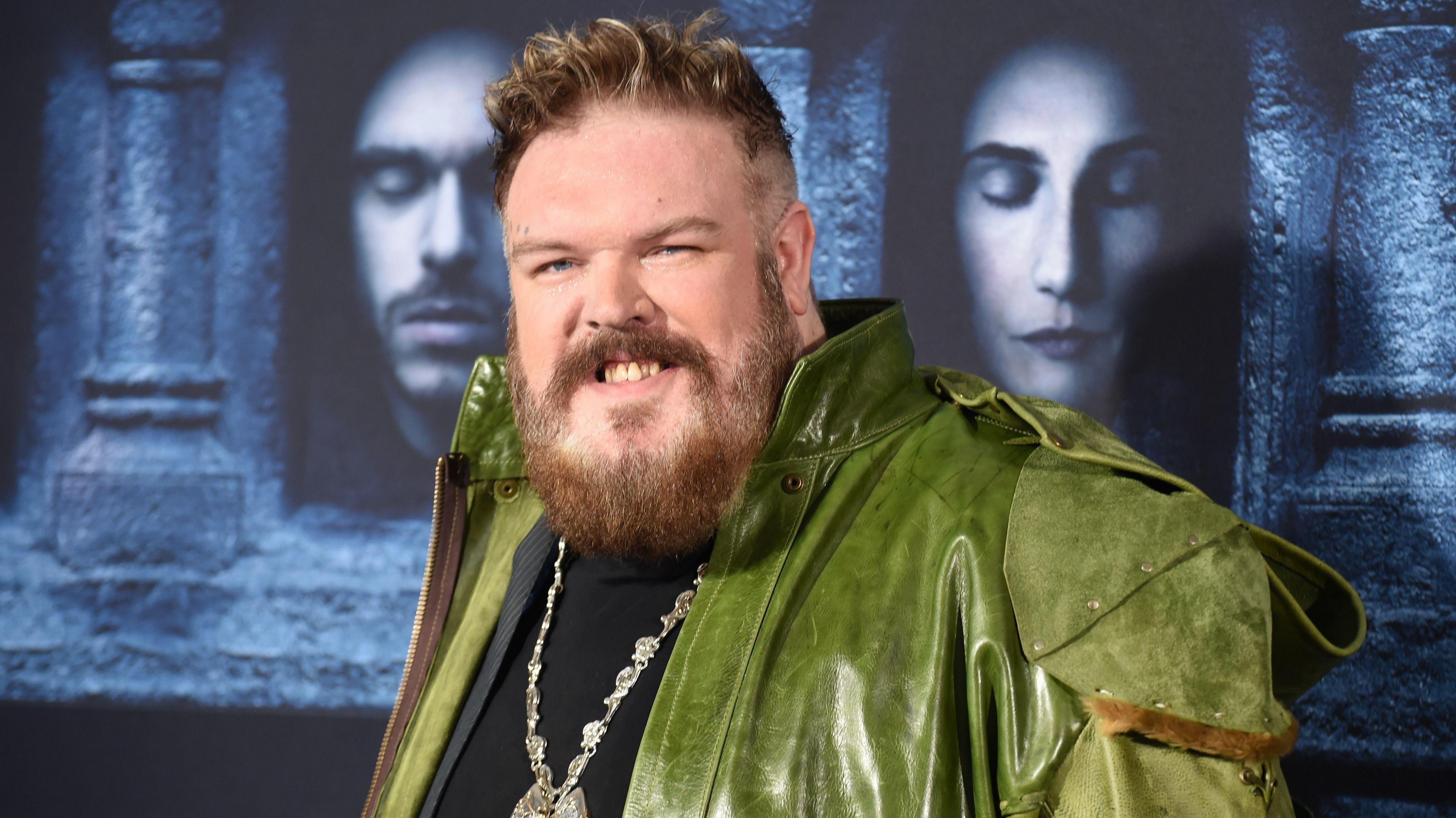 Kristian Nairn wearing a green leather jacket and medallion standing in front of Game of Thrones graphics at the premiere for the sixth season at TCL Chinese Theatre on April 10, 2016 in Hollywood