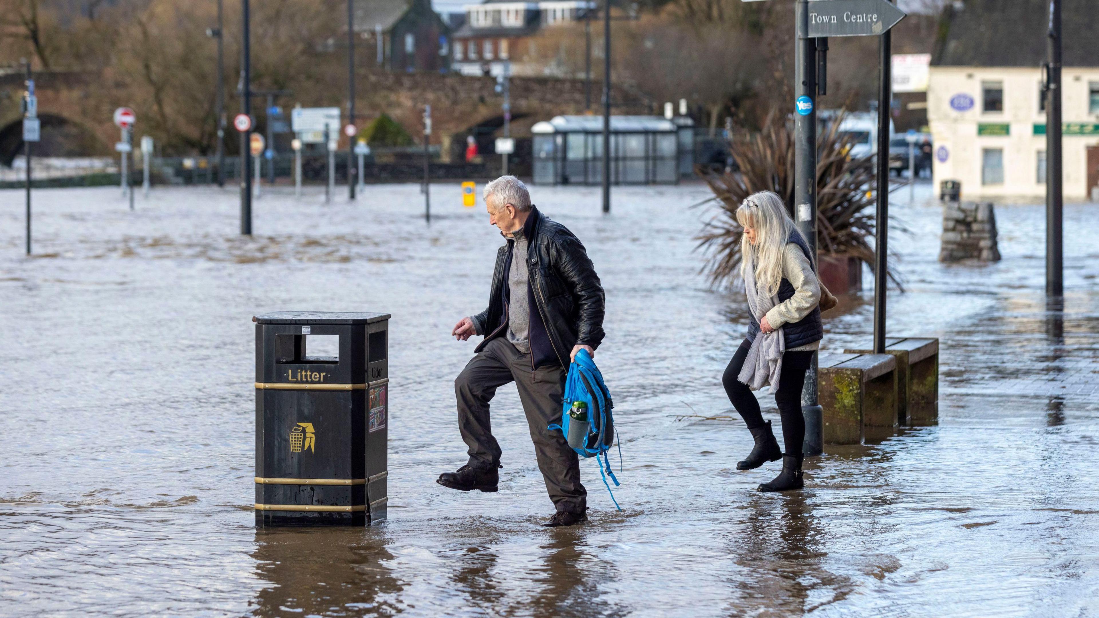 Two people - a man and a woman - walk through ankle deep water past a rubbish bin on the Whitesands in Dumfries