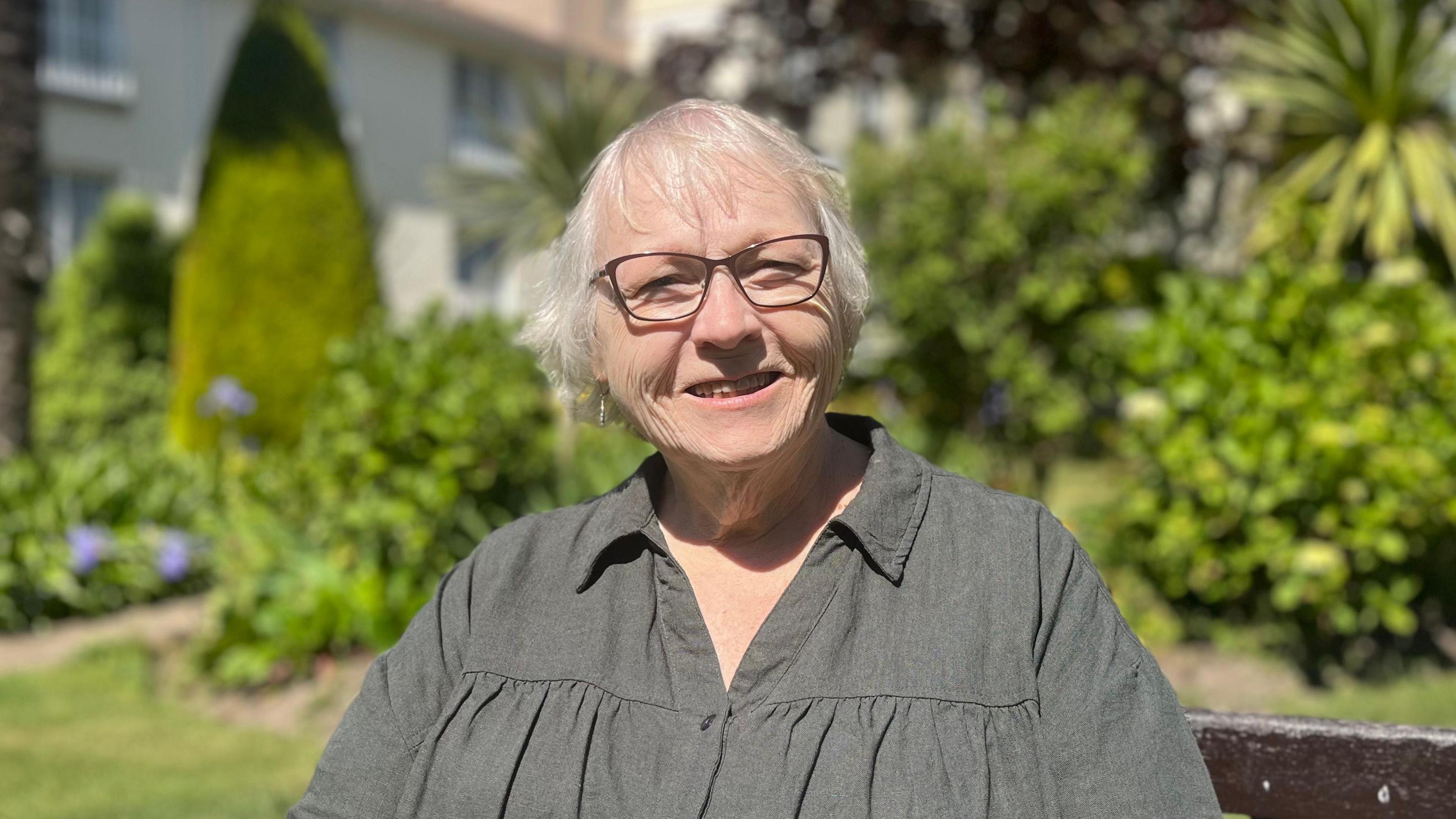 Jamie Poindexter is sat on a bench in a garden. She is wearing a dark green shirt and glasses and smiling into the camera.