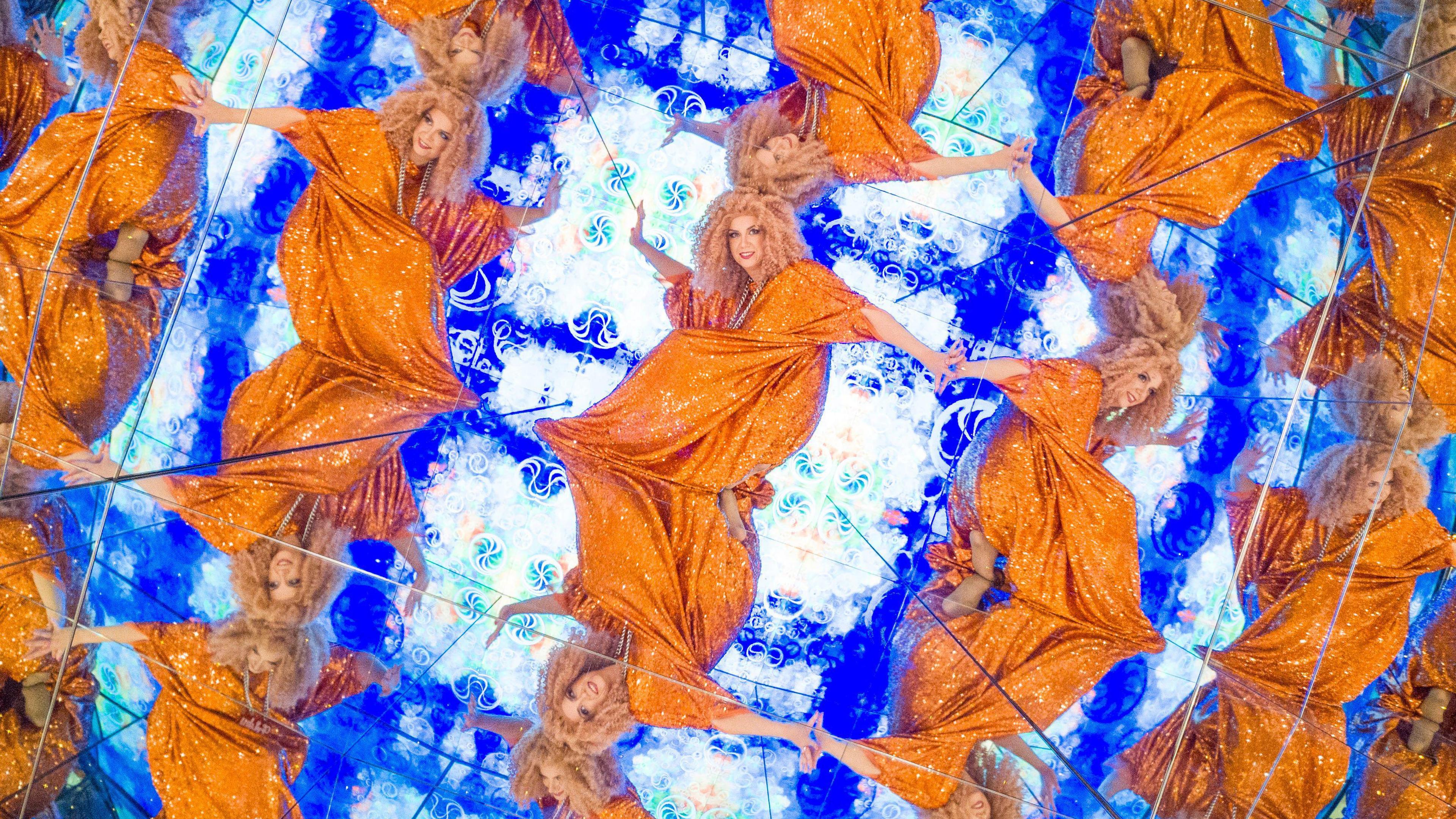 Miss Frisky - a blonde curly-haired woman, crouches inside a mirror box, reflecting her bright orange sequin dress onto dozens of mirrors around her.