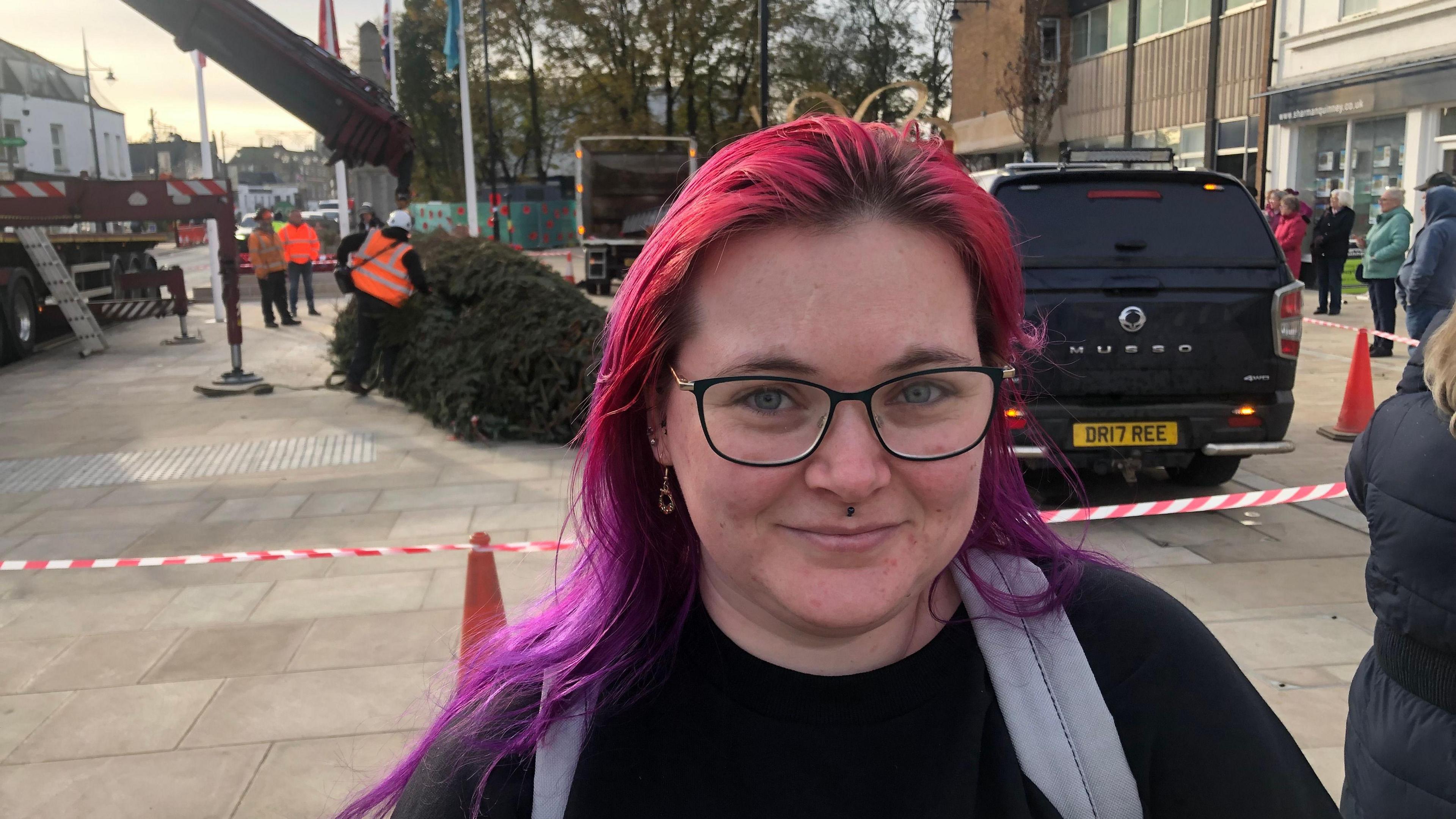 Emily Parkes has long red and purple hair over her shoulders. Her rucksack straps are visible. She has a piercing under her nose and is wearing spectacles. Behind her is a taped-off area where workmen are shaping the bottom of the trunk of the Christmas tree, which is lying on the pavement