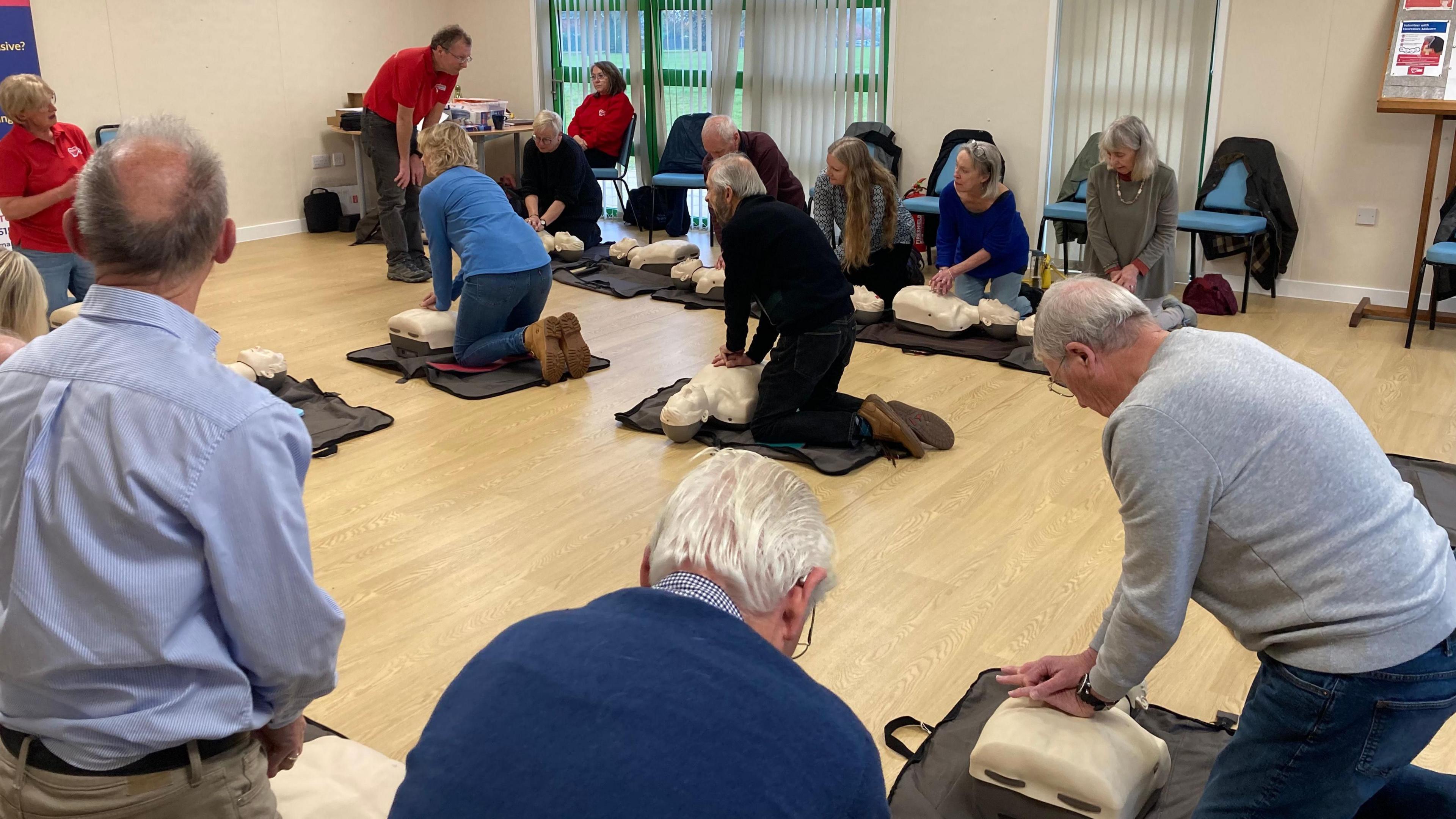 Ten people in a community centre room are all bent over practicing CPR on plastic dummies