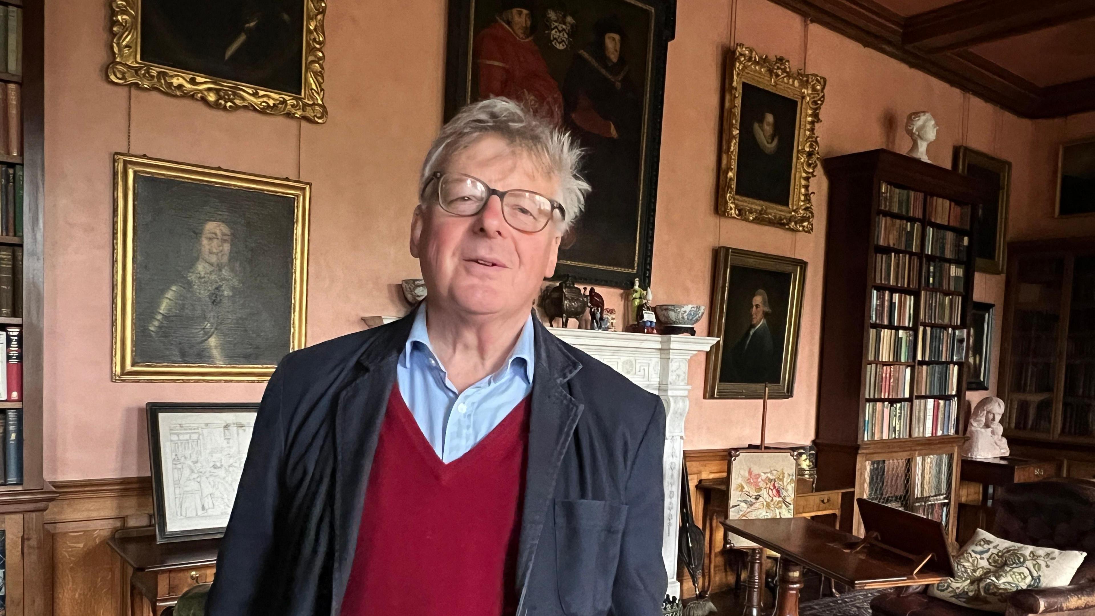 Richard Fletcher-Vane, the 2nd Baron Inglewood, inside his home. He has grey hair and spectacles and is wearing a red V-neck jumper, a blue shirt and a dark blazer jacket. Portraits are displayed in the background around a fireplace and a large bookcase stands to the right.