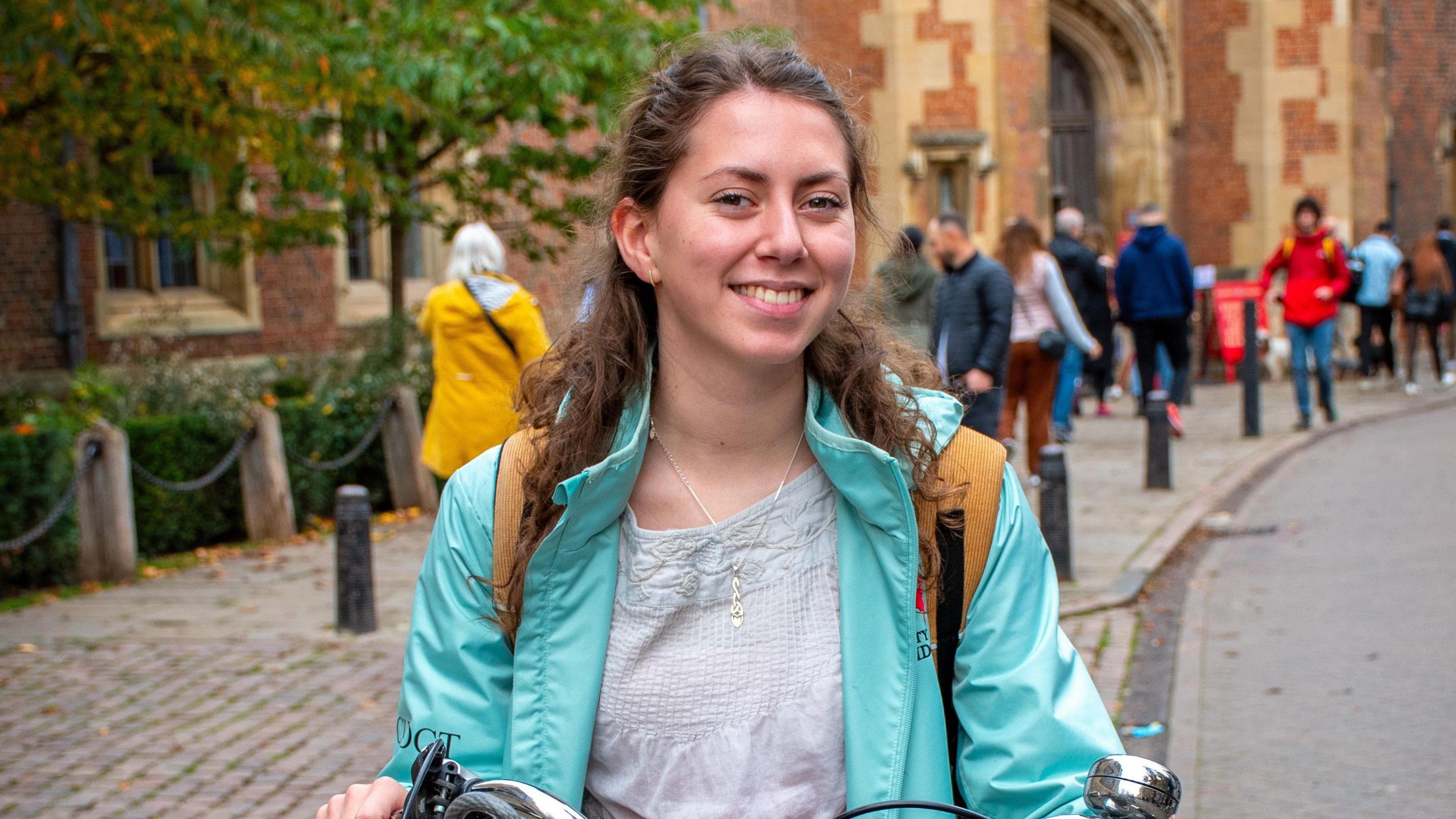 Gwennan yn sefyll o flaen coleg yng Nghaergrawnt. Mae hi'n gwisgo cot las ac yn dal beic.