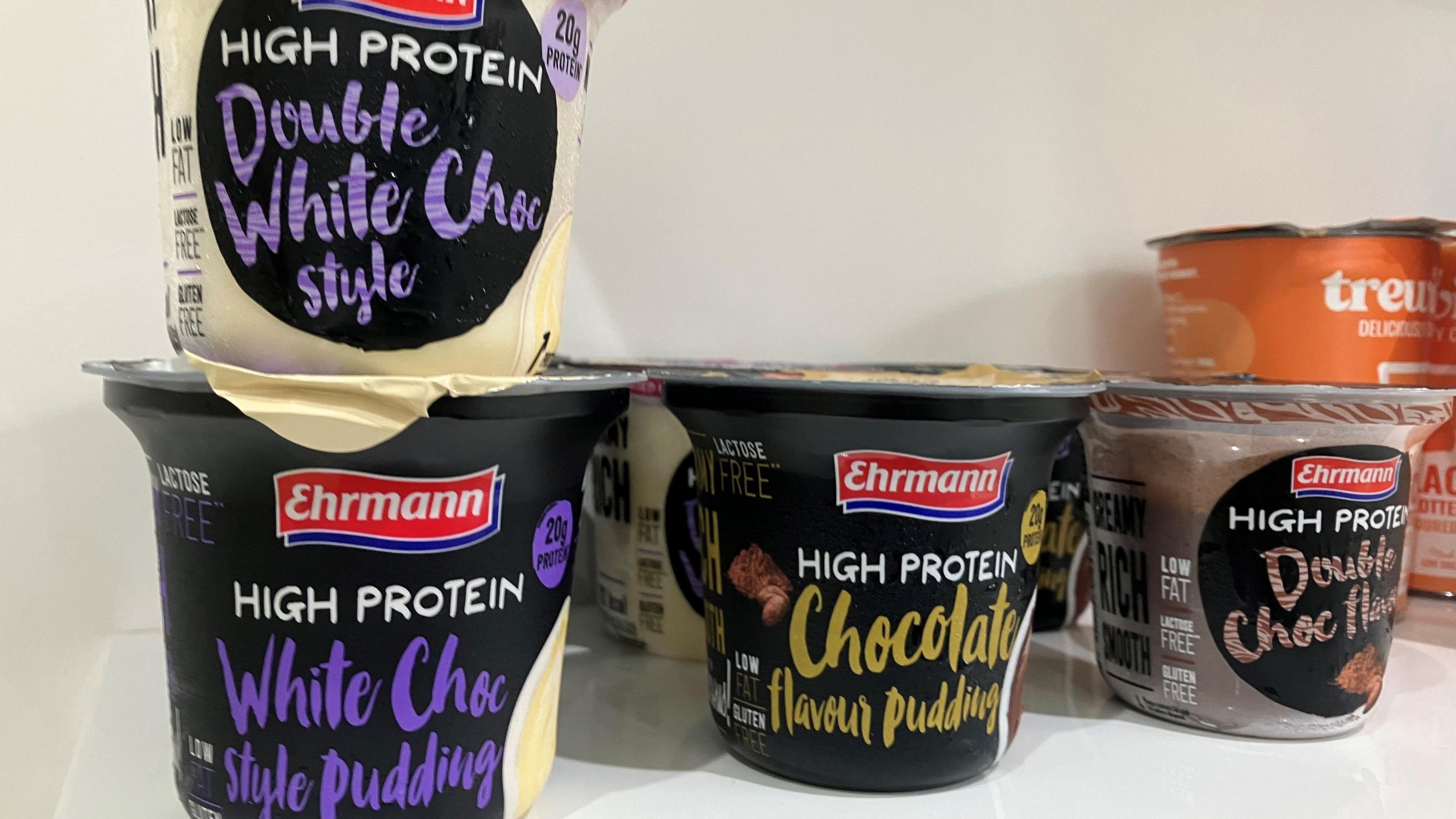 Pots lined up on a shelf of Ehrmann High Protein White Choc Style Pudding, Ehrmann High Protein Chocolate Flavour pudding, Ehrmann High Protein Double Choc flavour and Ehrmann High Protein Double White Choc Style.