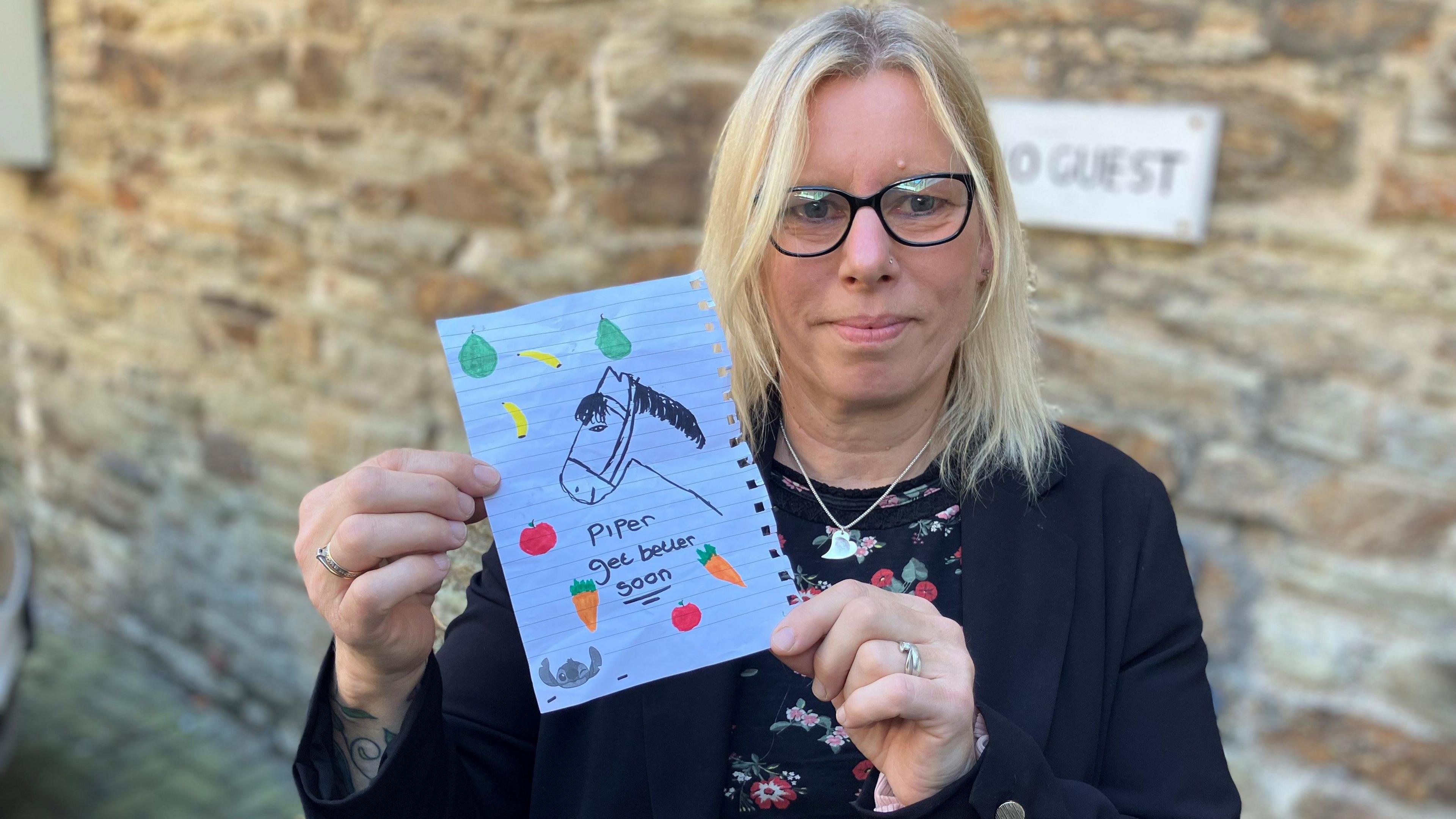 Woman holds up a drawing of a horse surrounded by fruits and vegetables with the words Piper get better soon