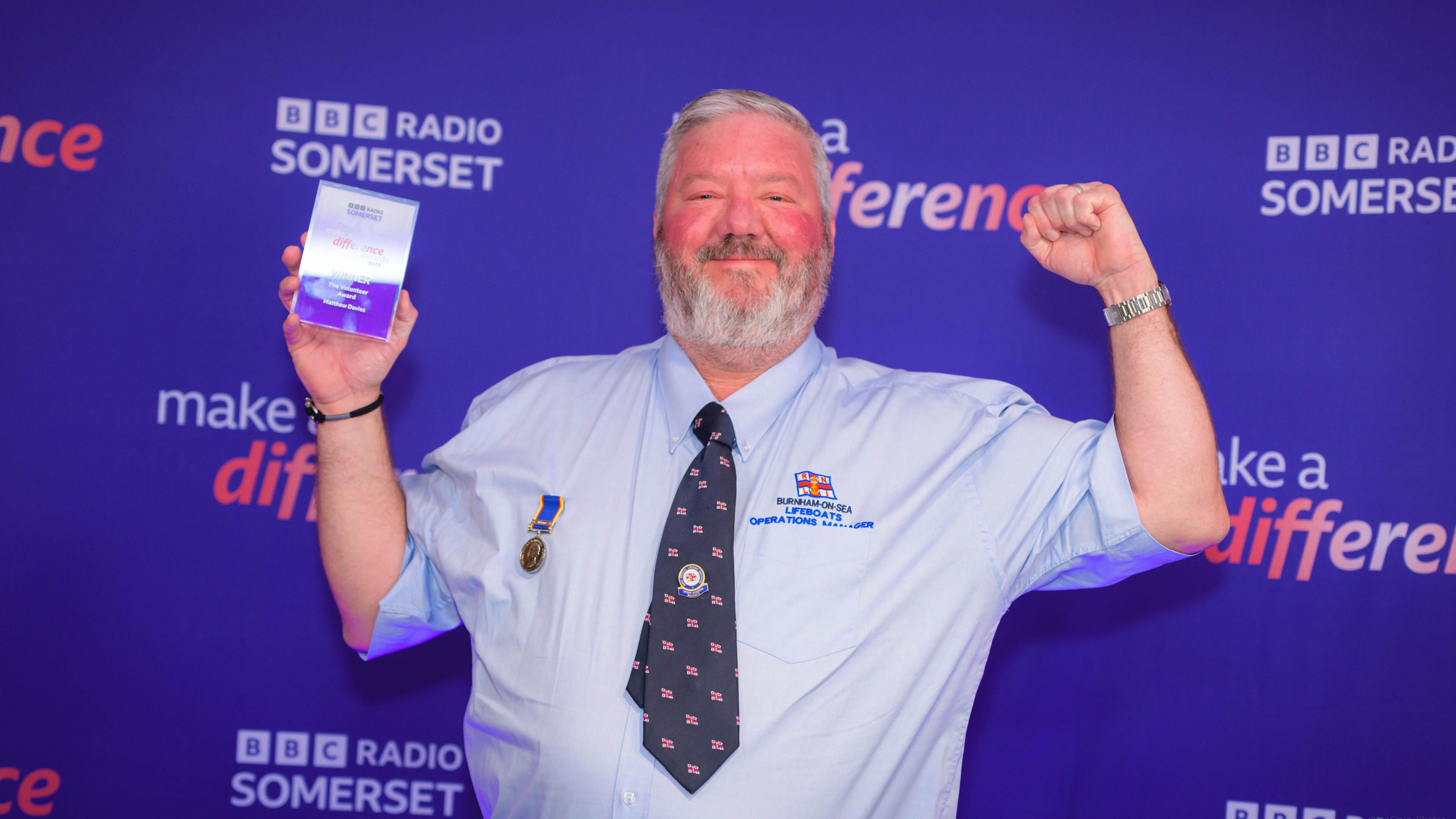 Man in blue shirt and tie cheers with his hands in the air whilst holding trophy in his left hand