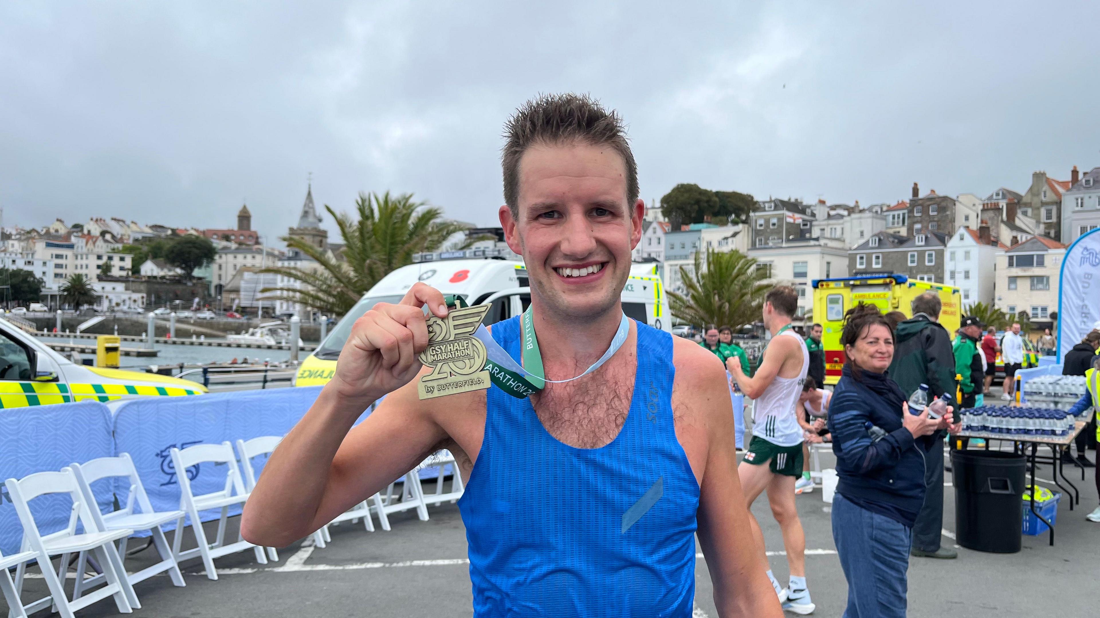 A smiling man in a blue running vest holds up a medal.