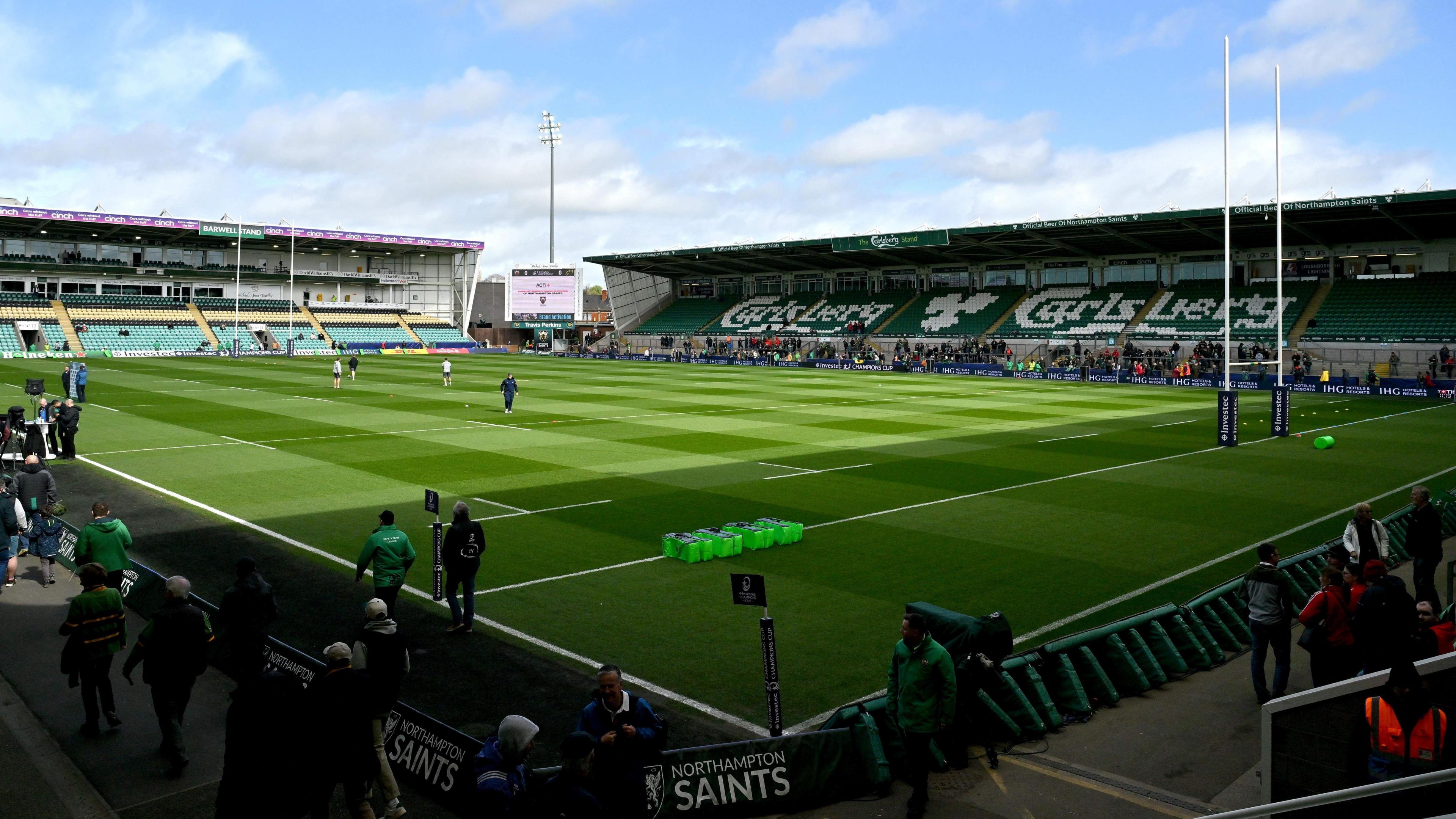 A photo from a corner of a rugby stadium. The sun and sky are clear and the pitch is bright green. There are two empty stands overlooking the pitch.