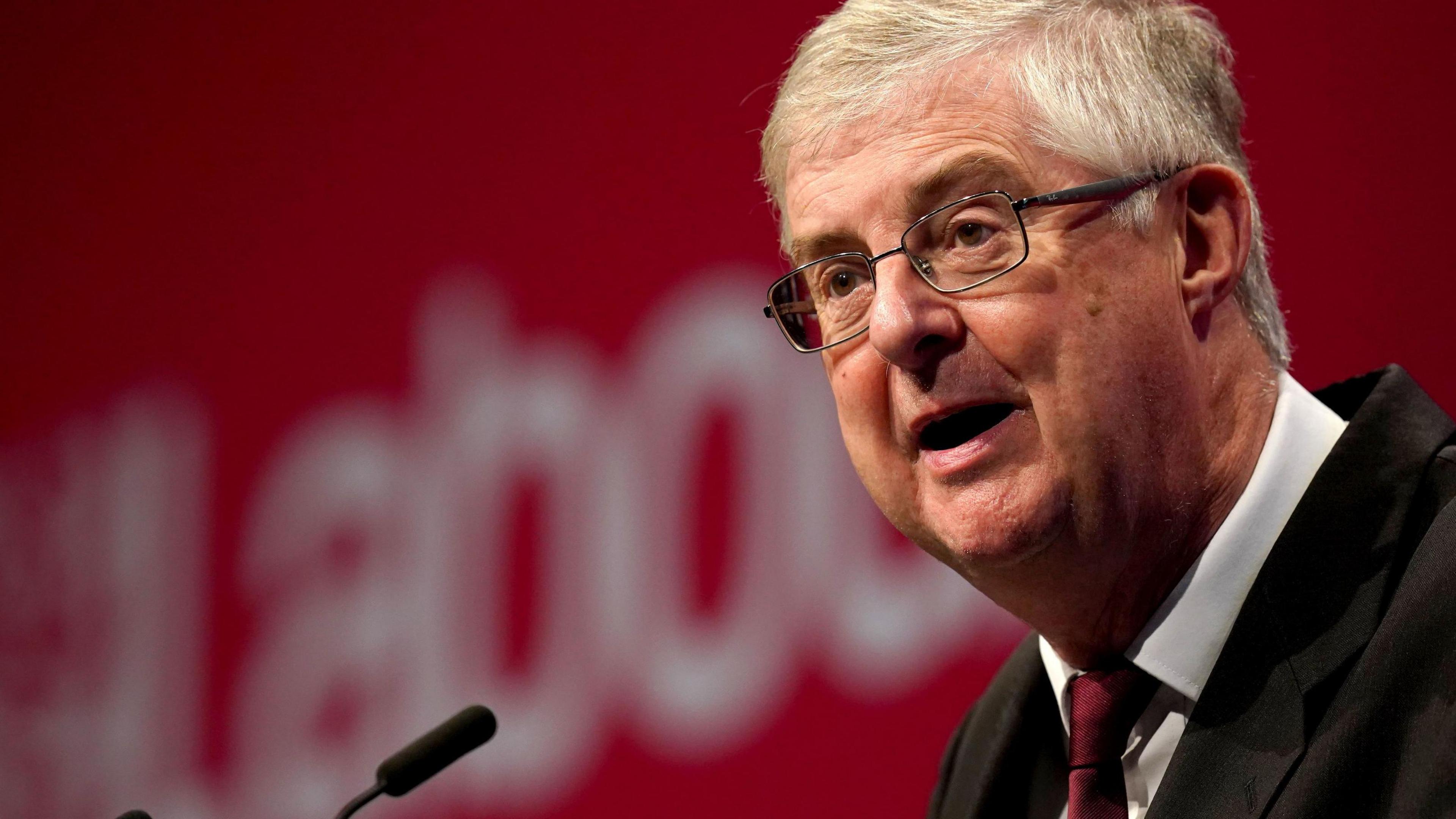 Llun o Mark Drakeford yn sefyll wrth podiwm. Mae e'n gwisgo siwt dywyll, crys gwyn a thei goch.
