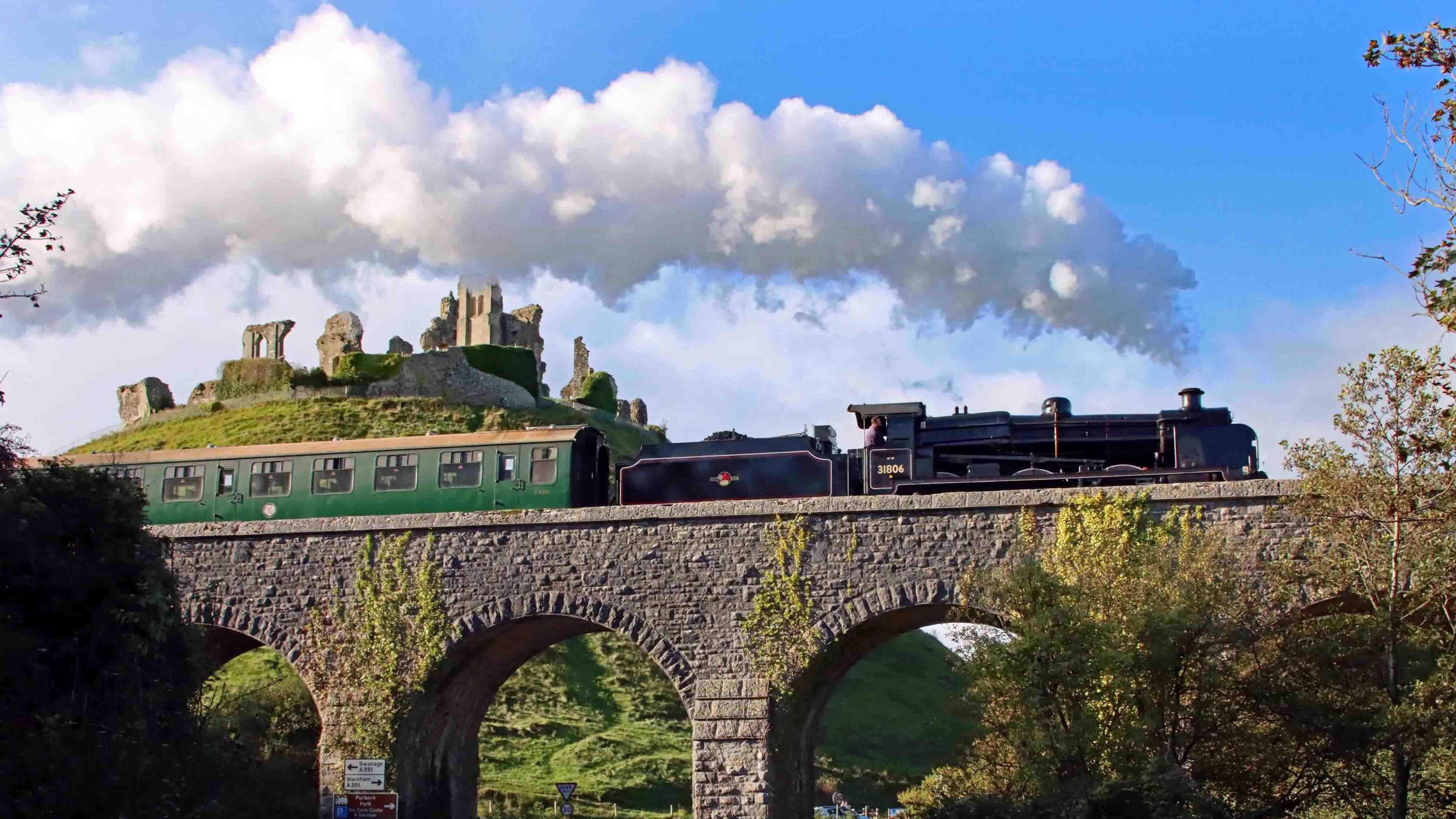 A black steam train moving over a railway bridge with the ruins of Corfe Castle on a hill behind it.