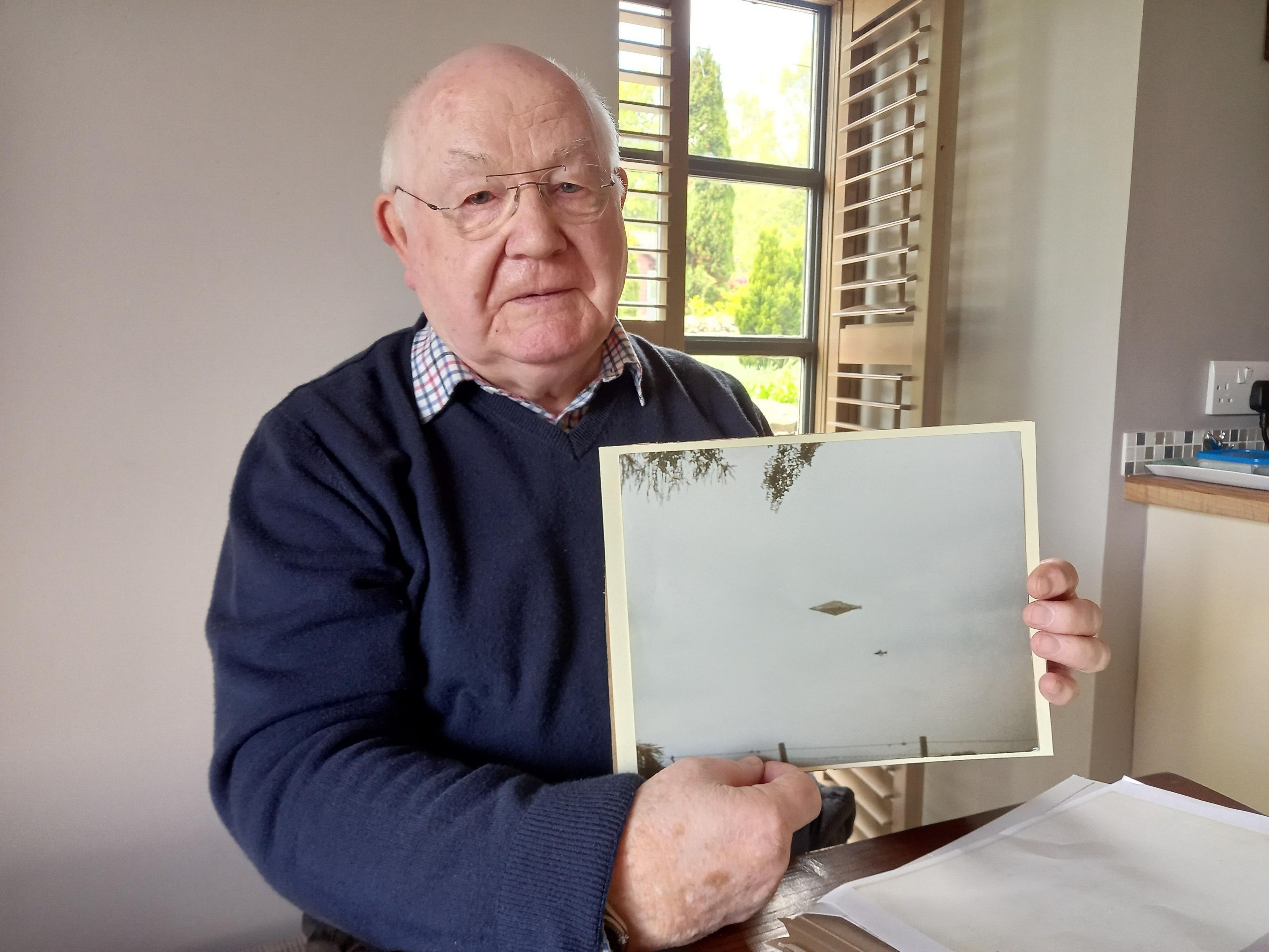 Craig Lindsay, an elderly man in a blue jumper and checked shirt, is holding up a copy of the Calvine photo. He appears to be sitting in a beige-coloured kitchen.