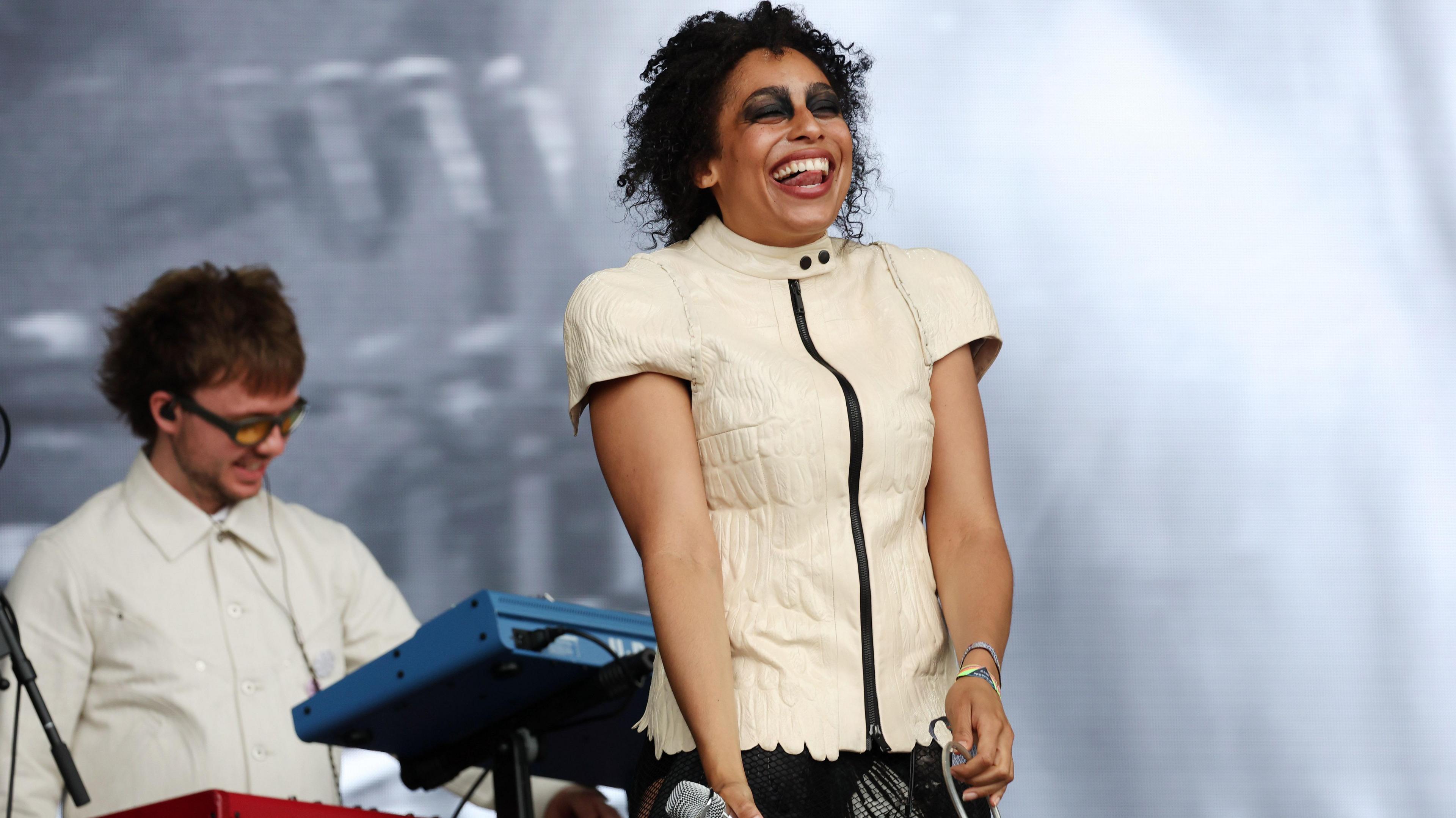 A woman is standing on stage in a white top laughing, with her tongue sticking out between her teeth. She has dark, black eyeshadow all around her eyes. Behind her is a man playing synths.