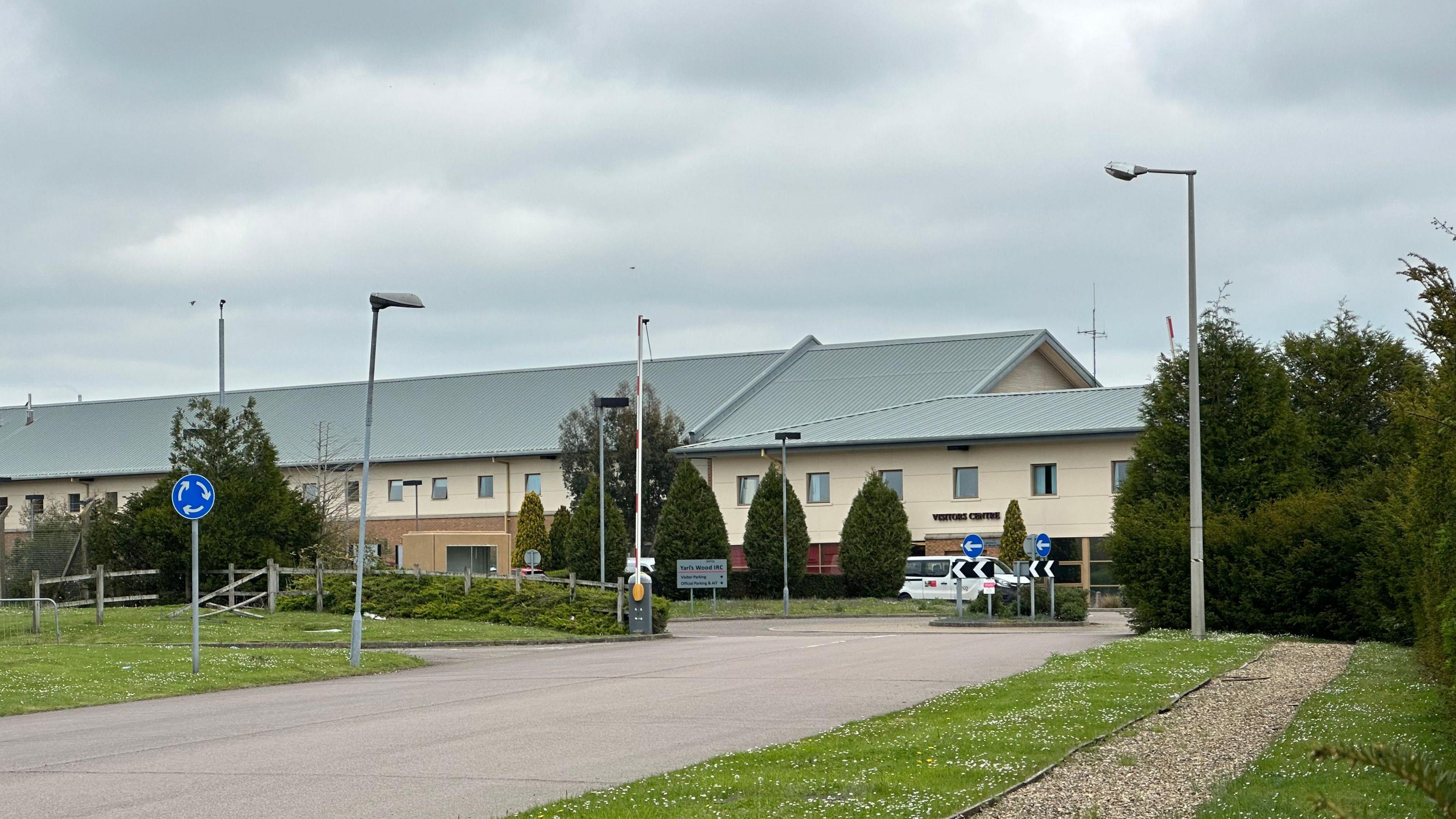 A general view of the entrance to Yarl's Wood. It shows a road leading to a roundabout ahead of the building. Signs can be seen to direct drivers and there is a raised gate allowing them inside.