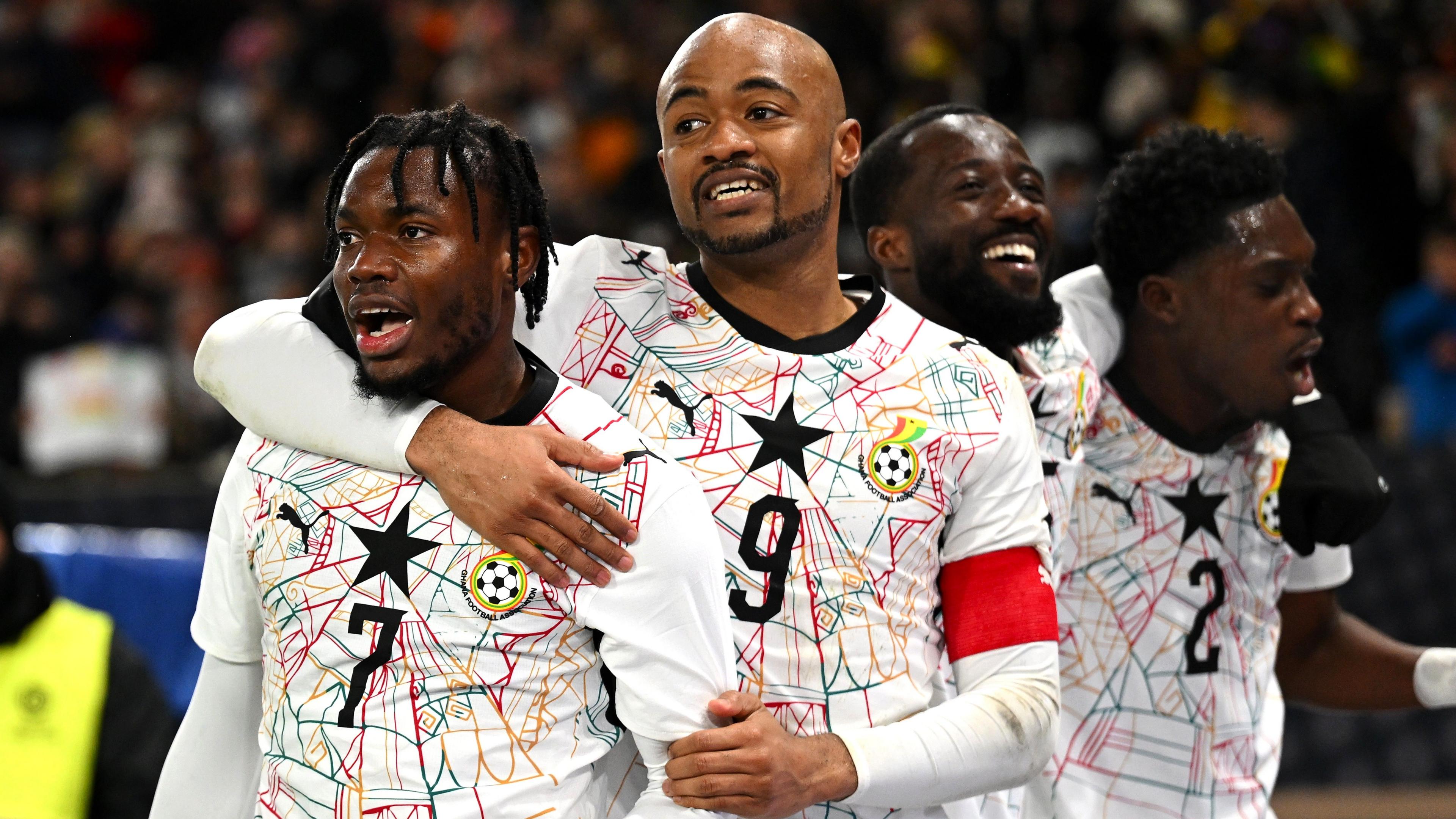 Abdul Fatawu celebrates scoring a goal with three team-mates during a match against Germany. Fatawu is on the left-hand side of the image while team-mate Jordan Ayew is next to him with his right arm wrapped around Fatawu's neck and his left arm clutching Fatawu's left arm. The two remaining players celebrate together behind the pair in the foreground. All four Ghanaians are visible from waist up and wear white Ghana home jerseys with an elaborate spiderweb design made up of thin threads in green, red and gold. The centre of the web is the black star which is in the centre of the players' chest. Under Fatawu's black star is a black number seven, while Ayew's shirt features a black number nine. Ayew also sports a red captain's armband on his left upper arm