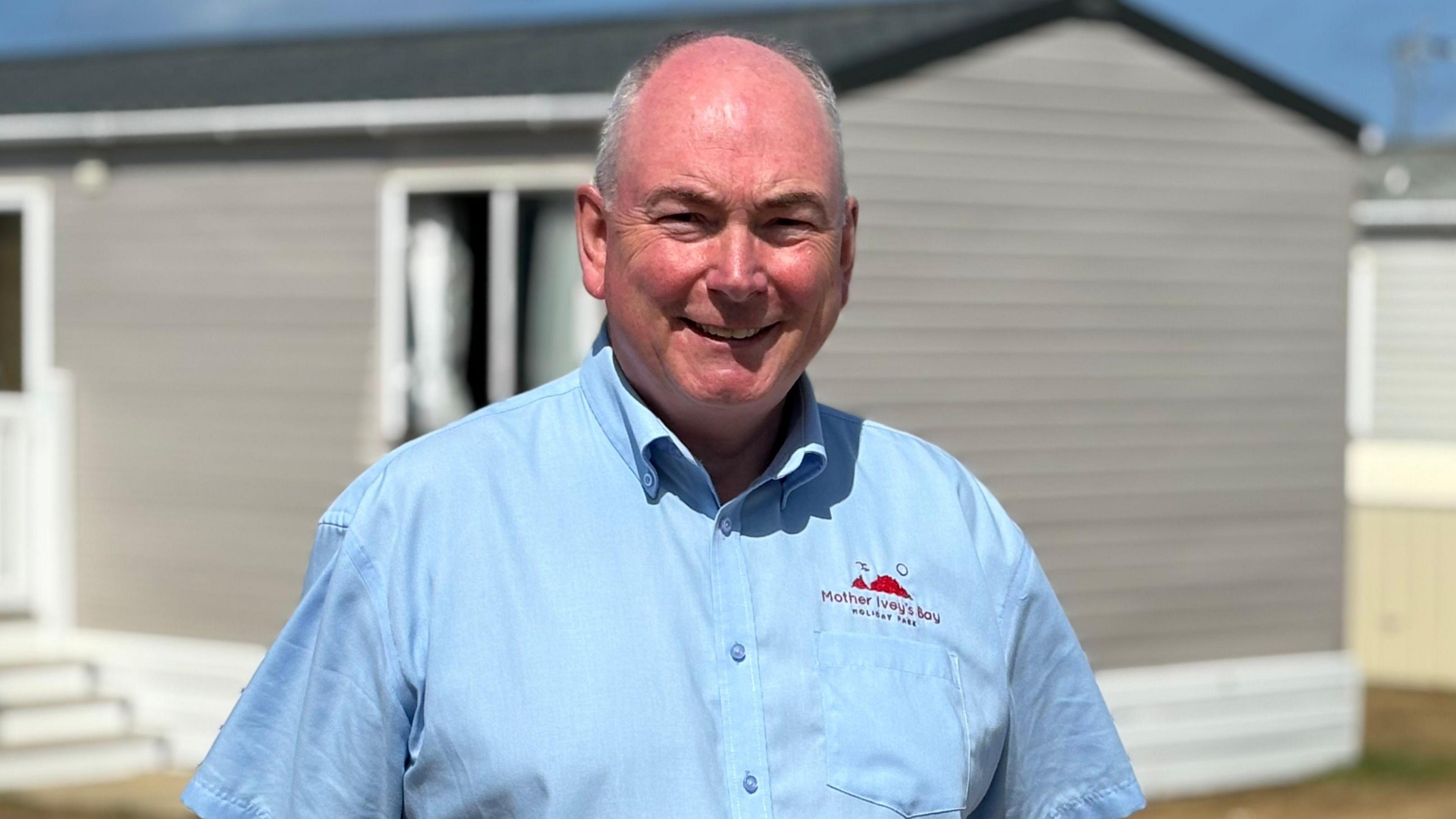 Patrick has grey hair and wears a light blue shirt - he stands with a static caravan behind him.
